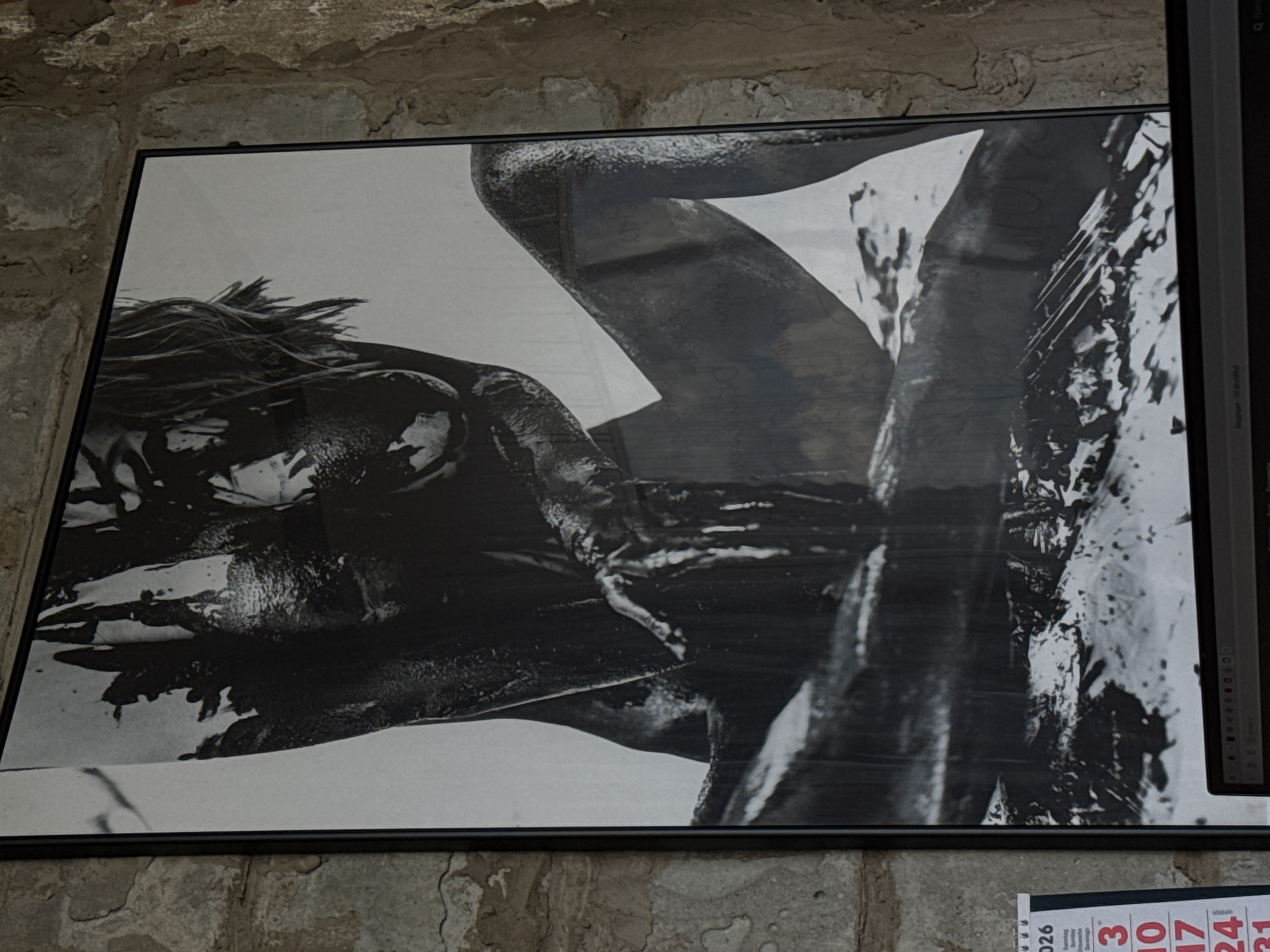 The image is a black and white photograph in a black frame, hanging on a roughly textured wall possibly made of stone or brick. The subject of the photograph appears to be a person covered in what looks like a dark, viscous substance, possibly mud or paint. The person's face is partially obscured by hair and the dark substance, but their shoulders, arms, and part of their torso are visible. The photograph is taken at an angle, and a calendar is partially visible at the bottom right corner, with numbers for the dates of March 2026. A portion of a computer monitor is visible on the right edge of the photograph. The location is Chaikovsky, Russia.