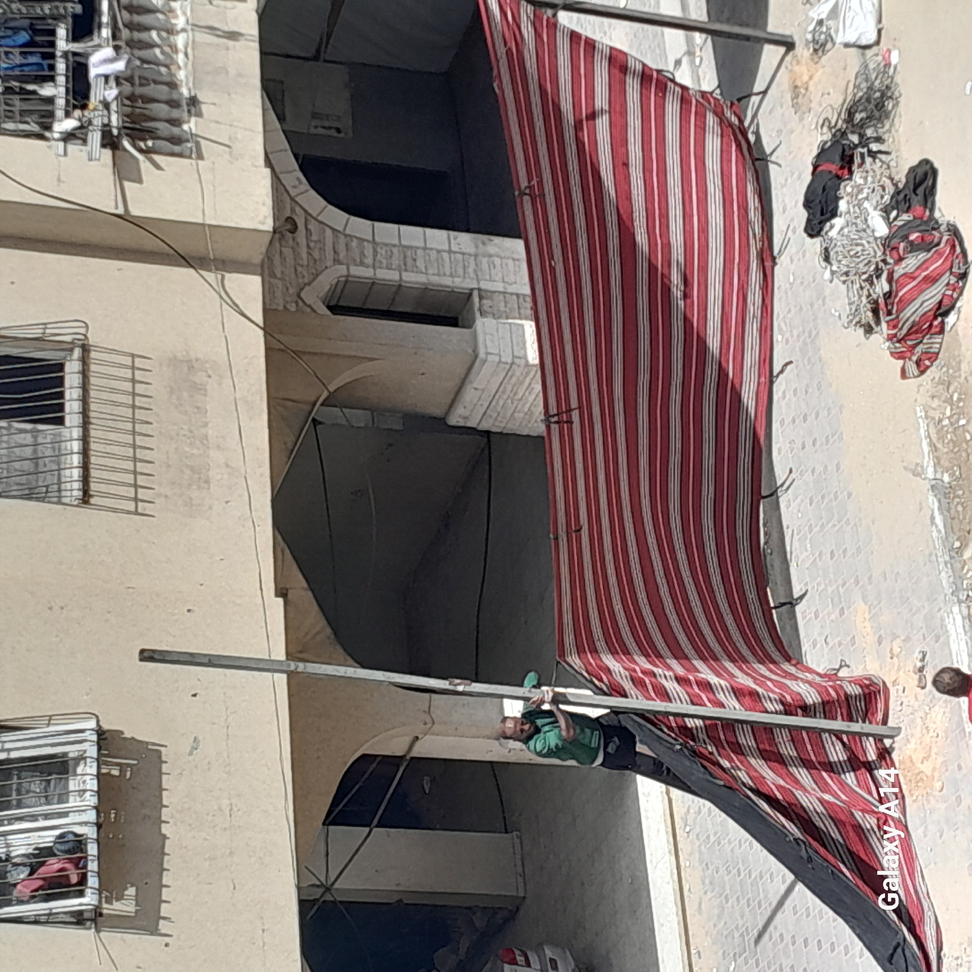 An overhead, slightly angled shot captures an outdoor scene, likely a street or alleyway adjacent to a multi-story building. The building features light-colored walls, windows secured with metal bars, and arched openings on lower levels.
A prominent red and white striped fabric awning extends diagonally across the frame, partially deployed or being adjusted. It is supported by two metal poles. A person, dressed in a green top and dark trousers, is visible interacting with one of these poles, appearing to be working on or around the awning structure. Their face is partially visible, and they seem to have a beard.
On the ground near the end of the awning, there are bundles of tangled ropes, fabrics, and other materials. A small, indistinct figure, possibly a child, is partially visible on the ground next to these items. Wires or cables run along the building's facade. Items, possibly laundry, are hanging outside an upper-floor window with security bars.
The scene appears to be taking place during the daytime, indicated by the clear lighting and visible shadows. The architectural style suggests an urban or semi-urban environment, possibly in a region with warm climates. The text "Galaxy A14" is watermarked on the striped fabric in the lower right portion of the image.