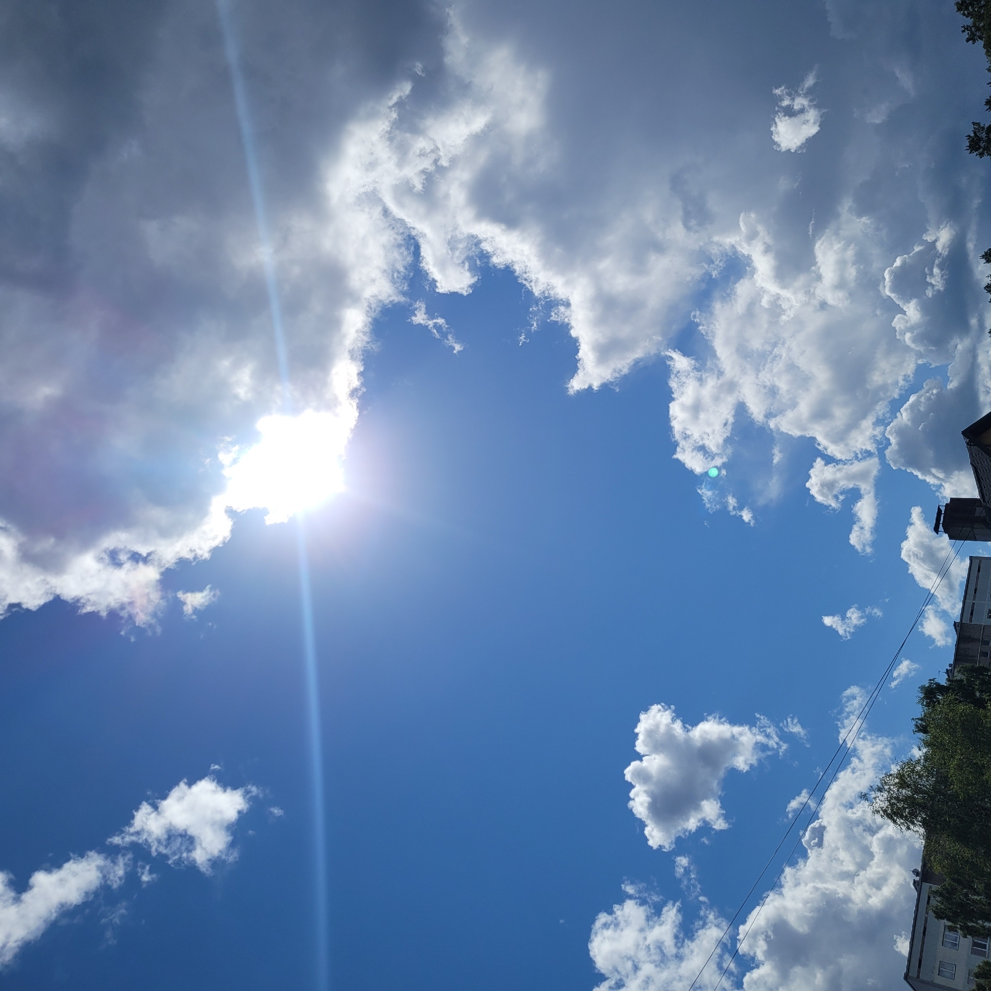 The image captures a bright, partly cloudy sky over a city. The sun is prominently visible, partially obscured by fluffy white clouds, creating a lens flare effect that streaks across the frame. In the lower portion of the image, the tops of buildings and trees are visible, suggesting an urban environment. Specifically, the scene is located in Jūrmala, Latvia. The sky transitions from a deep blue on the left to darker, more overcast clouds on the right, indicating a dynamic weather system. No people or specific actions are depicted. The environment appears to be outdoors during the daytime.