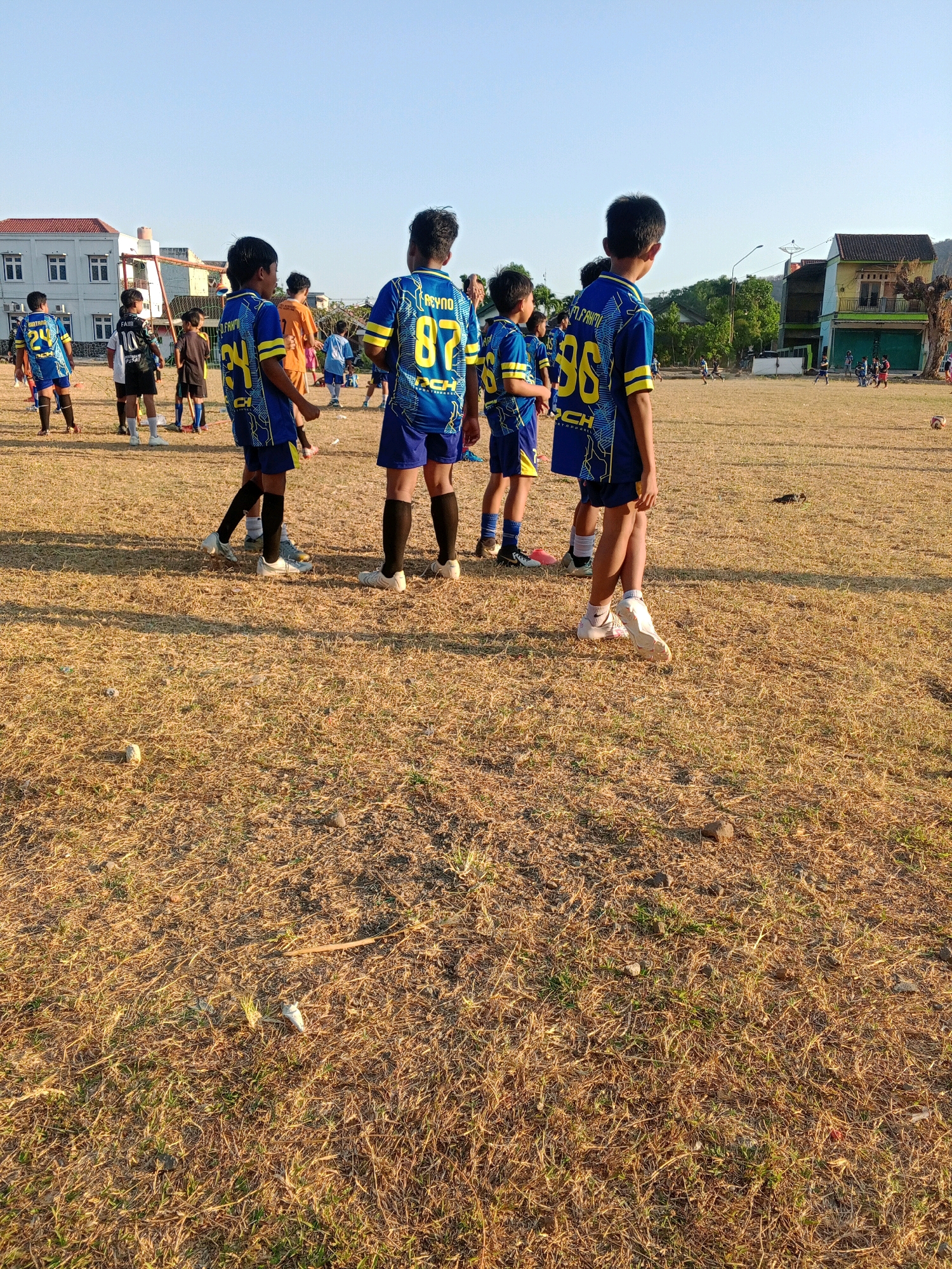 The image depicts a group of young boys in soccer uniforms on a dry, grassy field. The boys are wearing blue and yellow jerseys with numbers and names on the back. Some of them are standing and looking in different directions, while others appear to be in motion. In the background, there are buildings, trees, and a clear sky, suggesting an outdoor setting. The overall scene appears to be a soccer practice or game in the city of Giritirto, Indonesia, given the characteristic architecture and vegetation visible. The field itself is sparsely covered with dry grass and scattered with small stones and debris.