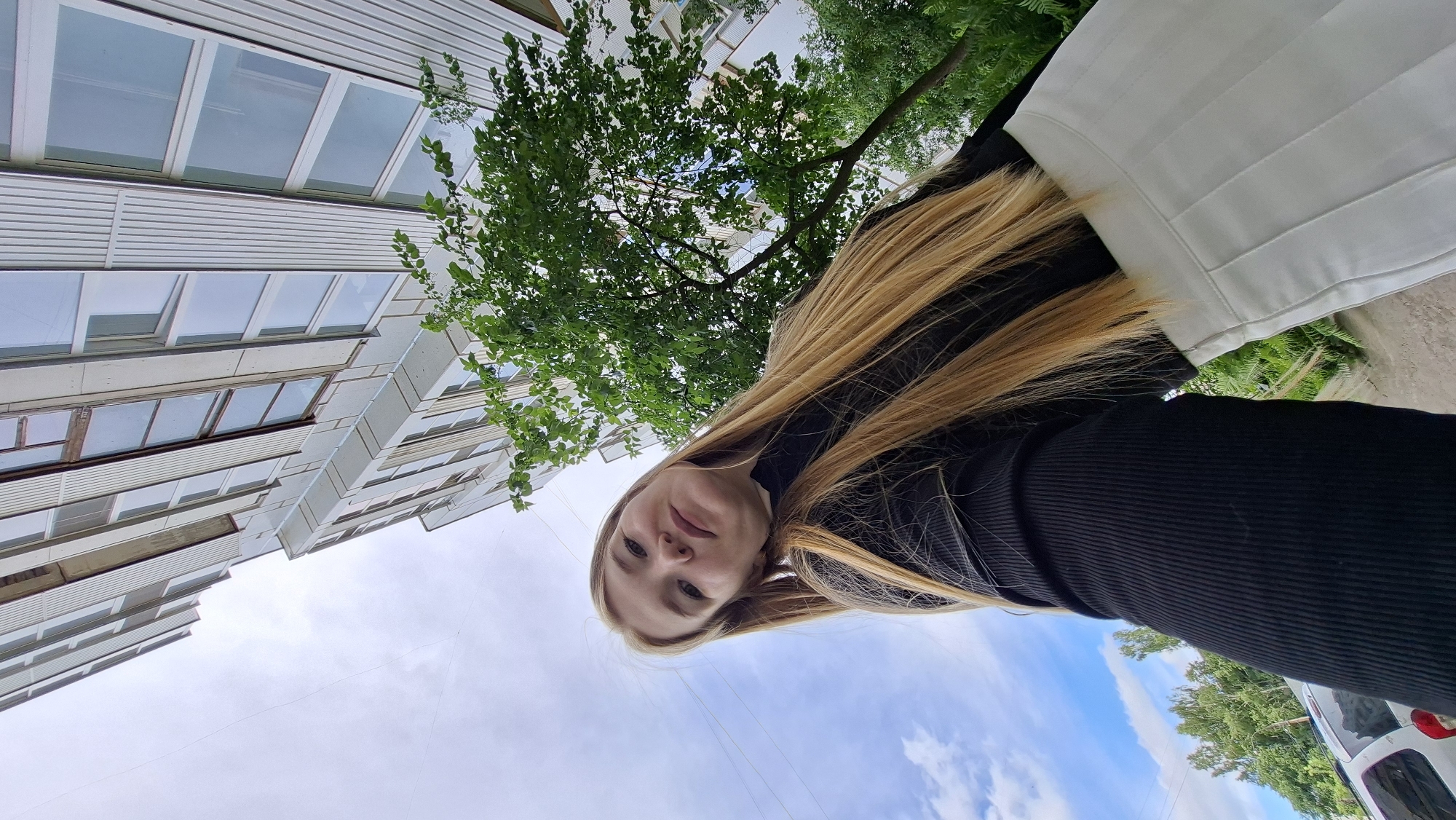 A young Caucasian woman with long blonde hair, wearing a black turtleneck and a white pleated skirt, poses for a low-angle selfie in front of a modern, multi-story building in Chaikovsky, Russia. The building features a white facade with rows of windows, and a lush green tree is partially visible in the frame, casting shadows. The sky is partly cloudy, with patches of blue and white clouds. In the background, the upper portion of a white car and some green foliage can be seen.