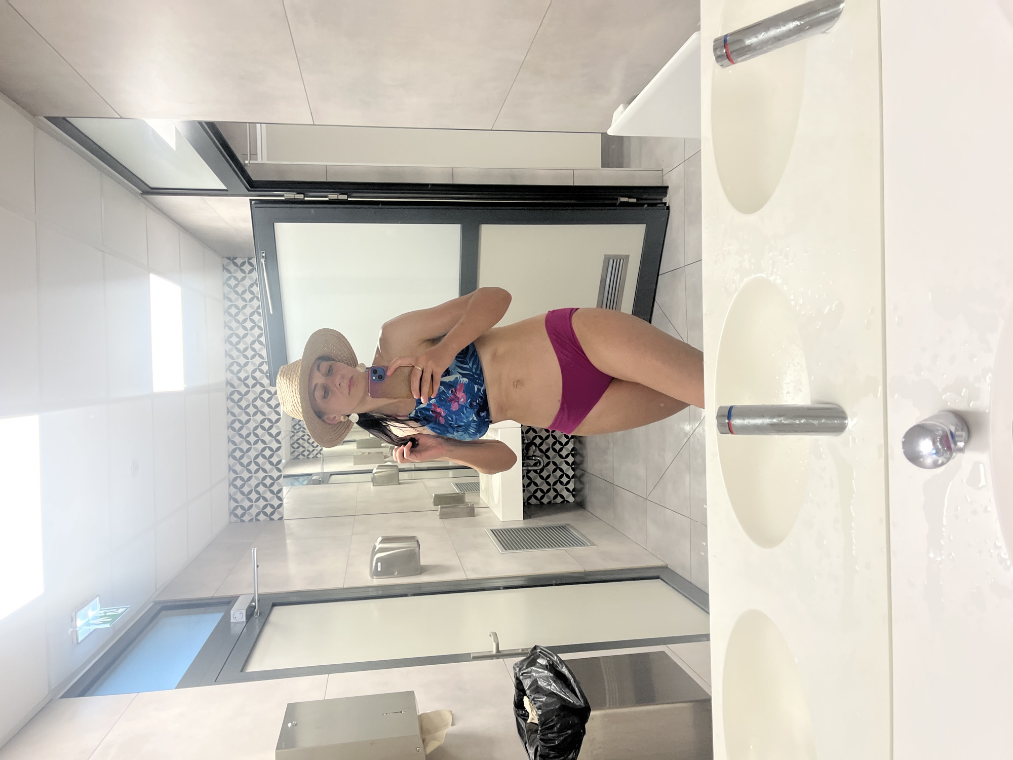 A woman is taking a mirror selfie in a public restroom. She is wearing a straw hat, a blue and pink floral bikini top, and magenta bikini bottoms. She is holding her phone in her right hand, and her left hand is touching her hair. The restroom is clean and modern, with light-colored tile walls and floors. There are several sinks with chrome faucets in the foreground, and a trash can is visible on the left. In the background, there are doors to stalls and a mirror that reflects the woman. The lighting is bright and even.