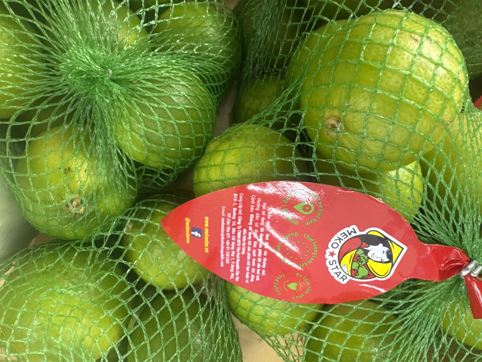 This media file is a close-up shot of several limes packaged in green mesh bags. The limes are a vibrant green color and appear to be fresh. A red, leaf-shaped label is attached to one of the bags, providing product information and branding. The label features text in Vietnamese, including brand names like "MEKO STAR" and "The Fruit Republic." It also includes phrases related to quality, safety, and origin, such as "Chất lượng cam kết" (Quality commitment) and "An toàn thực phẩm" (Food safety). The setting appears to be a retail or agricultural environment, likely a market or farm stand in Vietnam, given the language on the label and the "Viet Nam" text. There are no people or animals visible in the image, and the lighting suggests an indoor or shaded outdoor setting, as there are no strong shadows or indication of a specific time of day. The focus is entirely on the produce and its packaging.