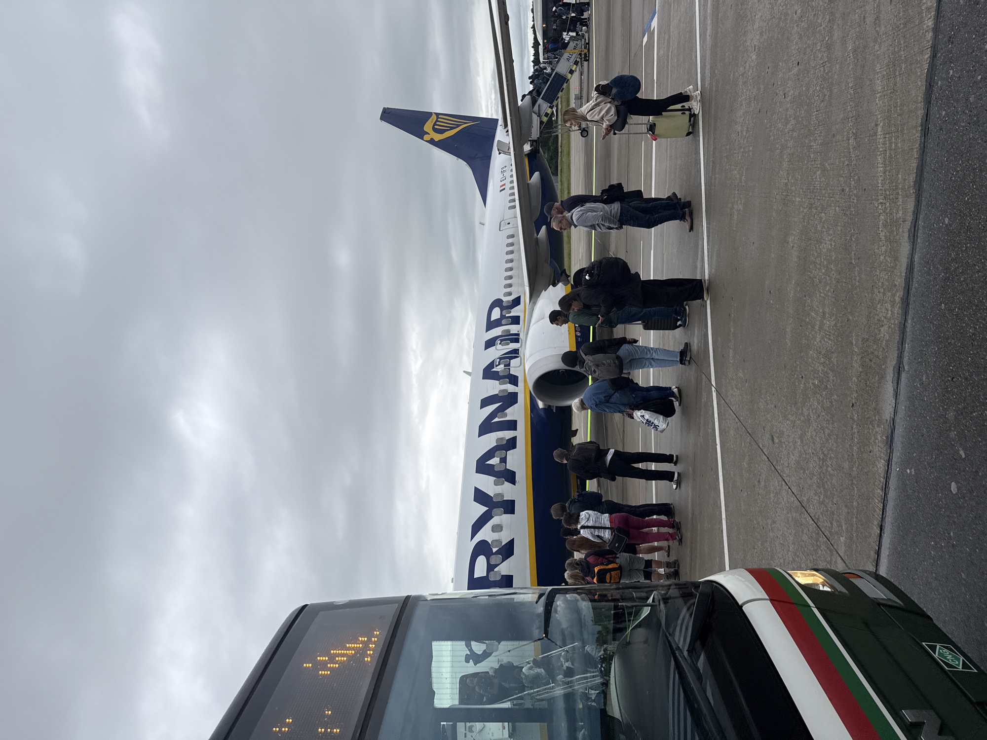The image displays an active airport tarmac scene, likely in Künersberg, Germany, under an overcast sky. Numerous passengers are captured in the process of boarding a Ryanair airplane.
The central subject is a large white and blue Ryanair aircraft, identified by the prominent "RYANAIR" lettering along its fuselage and the distinctive harp logo on its tail. The airplane's registration number, "EI-EFS," is visible on the tail fin. Passengers are queuing in multiple lines, extending from a ground vehicle towards the aircraft. Some individuals are already ascending the mobile boarding stairs, which has the text "ALL ie" visible on its side, to enter the airplane. Many passengers are carrying carry-on luggage or wearing backpacks.
In the foreground, the front section of a bus is visible. This bus features a distinctive design with green, white, and red stripes along its side. It has a lit digital destination display on its front, though the specific text is unreadable, and a "CNG" sticker is present on its body. The ground is a typical airport tarmac, marked with white lines. The sky is uniformly gray and cloudy, indicating an overcast day. The overall atmosphere depicts the organized activity of passengers transferring from a bus to an aircraft for departure.