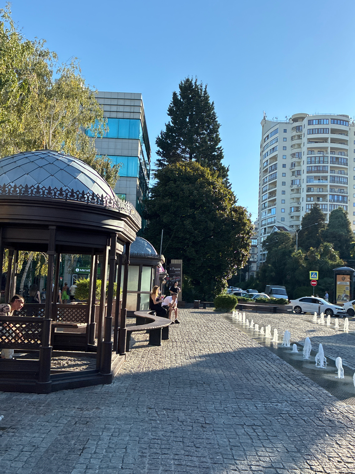 The image depicts an outdoor scene in Sochi, Russia, likely during daytime given the clear blue sky and the bright sunlight casting shadows. In the foreground, a decorative dark brown gazebo with a domed roof and intricate metalwork provides shade for several people. Benches are visible inside the gazebo, and some individuals are seated on them.
Beyond the gazebo, a paved walkway made of cobblestones leads towards the background. A row of small fountains sprays water onto the walkway, creating a decorative water feature. Several cars are parked along the street in the middle ground, with some moving.
The background features modern urban architecture, including a multi-story building with a gray facade and bright blue windows, and a taller, cream-colored residential building with balconies and numerous windows. Lush green trees, including a tall conifer, are interspersed between the buildings, adding a natural element to the urban landscape. A street sign indicating a "Market" is visible on the right side of the image. The overall impression is a well-maintained public space in a city with a mix of modern and traditional architectural styles.