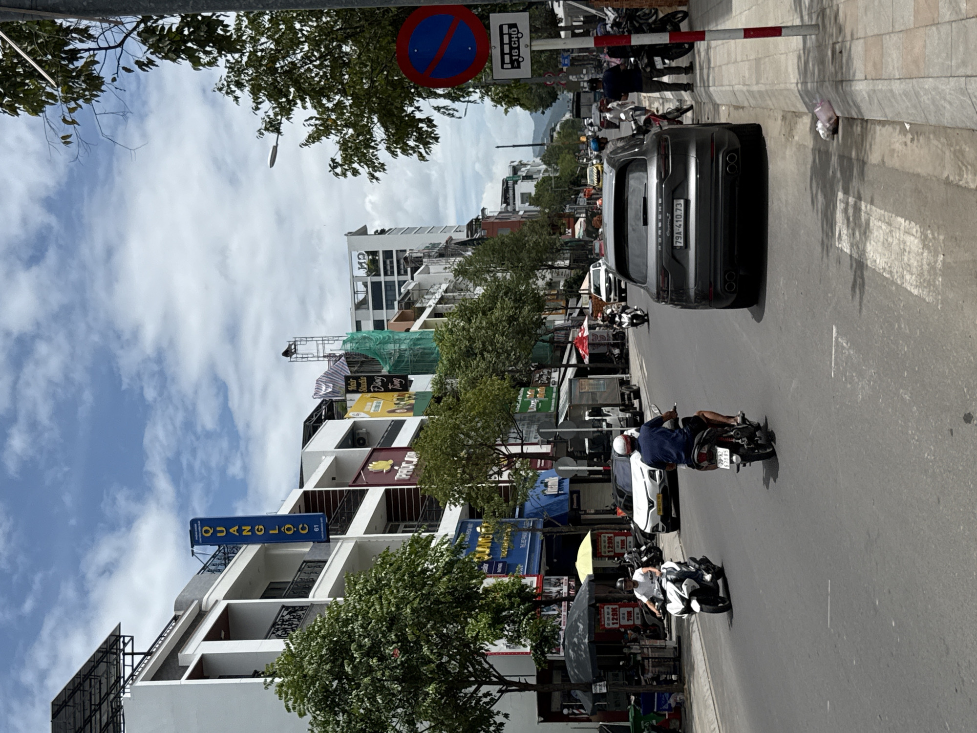 The image depicts a street scene in Nha Trang, Vietnam, on a cloudy day. The street is lined with multi-story buildings housing various businesses, including a "QUANGLOC" sign in blue and yellow. Several trees are planted along the sidewalk, casting shadows on the asphalt.
Traffic is present, with several motorbikes and a dark grey Porsche Cayenne visible. Two individuals are riding motorbikes in the foreground. One person on a white motorbike is moving away from the viewer, and another on a dark motorbike is closer and facing away. A white sedan is parked on the side of the road.
On the right side of the street, a red and blue "no stopping" traffic sign is mounted on a pole, accompanied by a sign indicating "16 CHO" (16 seats), likely a bus stop designation. Pedestrians are present on the sidewalk, and parked motorbikes are also visible. Construction appears to be underway on one of the buildings, with scaffolding and green netting. The overall environment suggests an urban street with moderate commercial activity.
