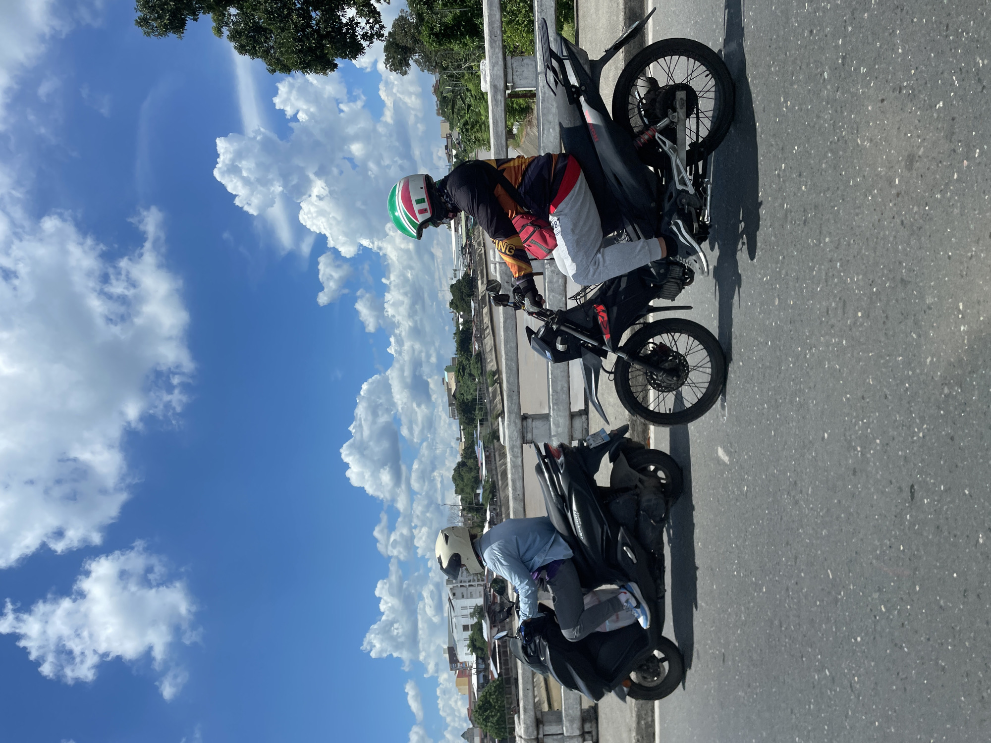 Two individuals are riding motorcycles on a bridge, with the Davao River visible below. The sky is blue with scattered white clouds. The person on the left is riding a dark-colored scooter and is wearing a light-colored helmet and a light blue long-sleeved shirt. The person on the right is riding a dirt bike style motorcycle and is wearing a green, white, and red helmet, a black and orange jacket, and light gray pants. They are both wearing face masks. The bridge has a concrete railing. In the background, there are buildings and trees on the riverbanks. The scene appears to be captured on a sunny day in Davao City, Philippines.