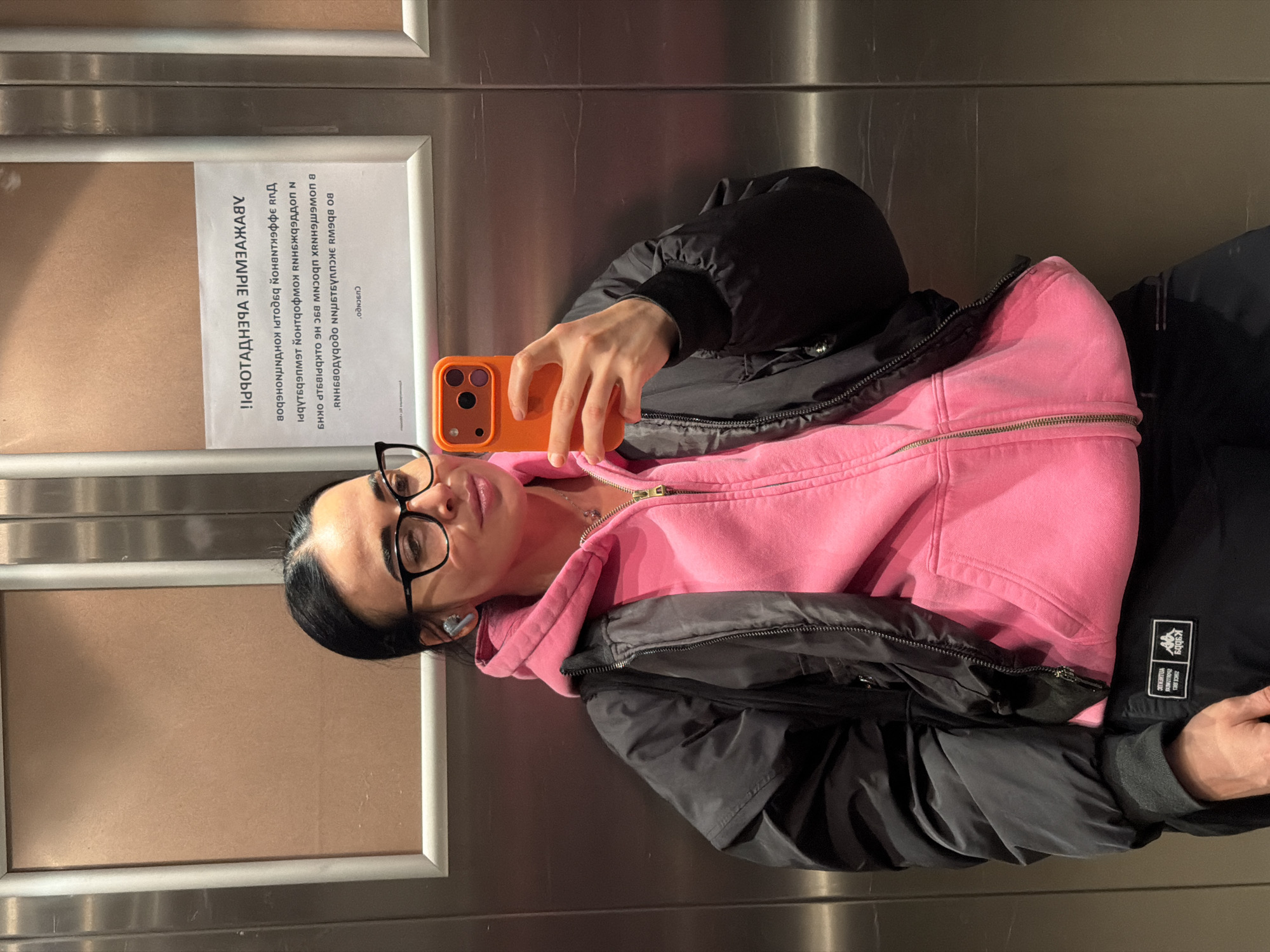 The image shows a woman with dark hair and glasses taking a selfie in what appears to be an elevator. She is wearing a pink hoodie and a black jacket. Her phone has an orange case. Behind her is a framed notice written in Russian. The notice contains text in the Russian language, mentioning what appears to be technical specifications. The visible text is "ОБЪЯВЛЕНИЕ УБЕНТДТАОРПI" and other text that is inverted. The woman's location in Saint Petersburg, Russia, is not explicitly indicated in the image.