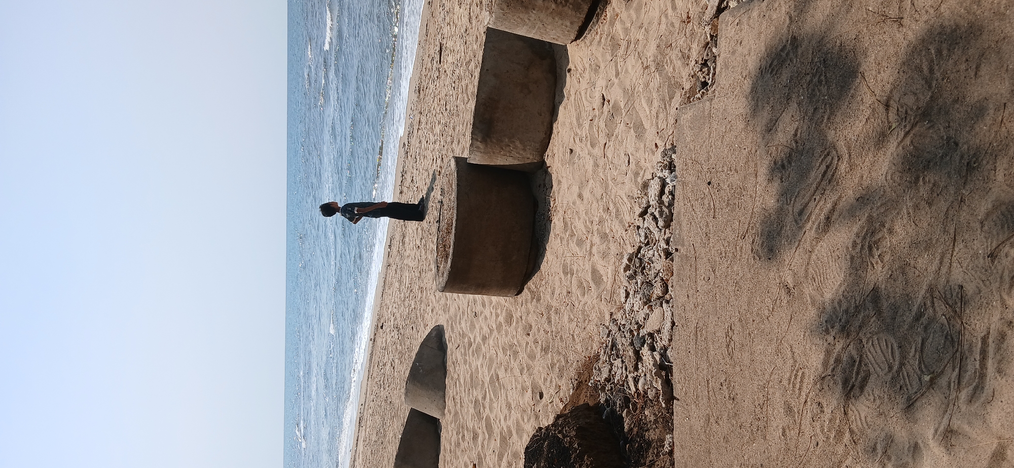 A lone figure, a child, stands on a sandy beach in Banyuglugur, Indonesia, facing the calm sea. The child is silhouetted against the water, wearing a dark shirt and pants. In the foreground, several large, concrete cylindrical structures are partially buried in the sand. The beach is covered in light brown sand, with some scattered debris and vegetation. The sea is a clear blue, with gentle waves lapping at the shore. The sky is a pale blue, with no clouds visible. The overall impression is one of quiet contemplation and a vast, open natural landscape.