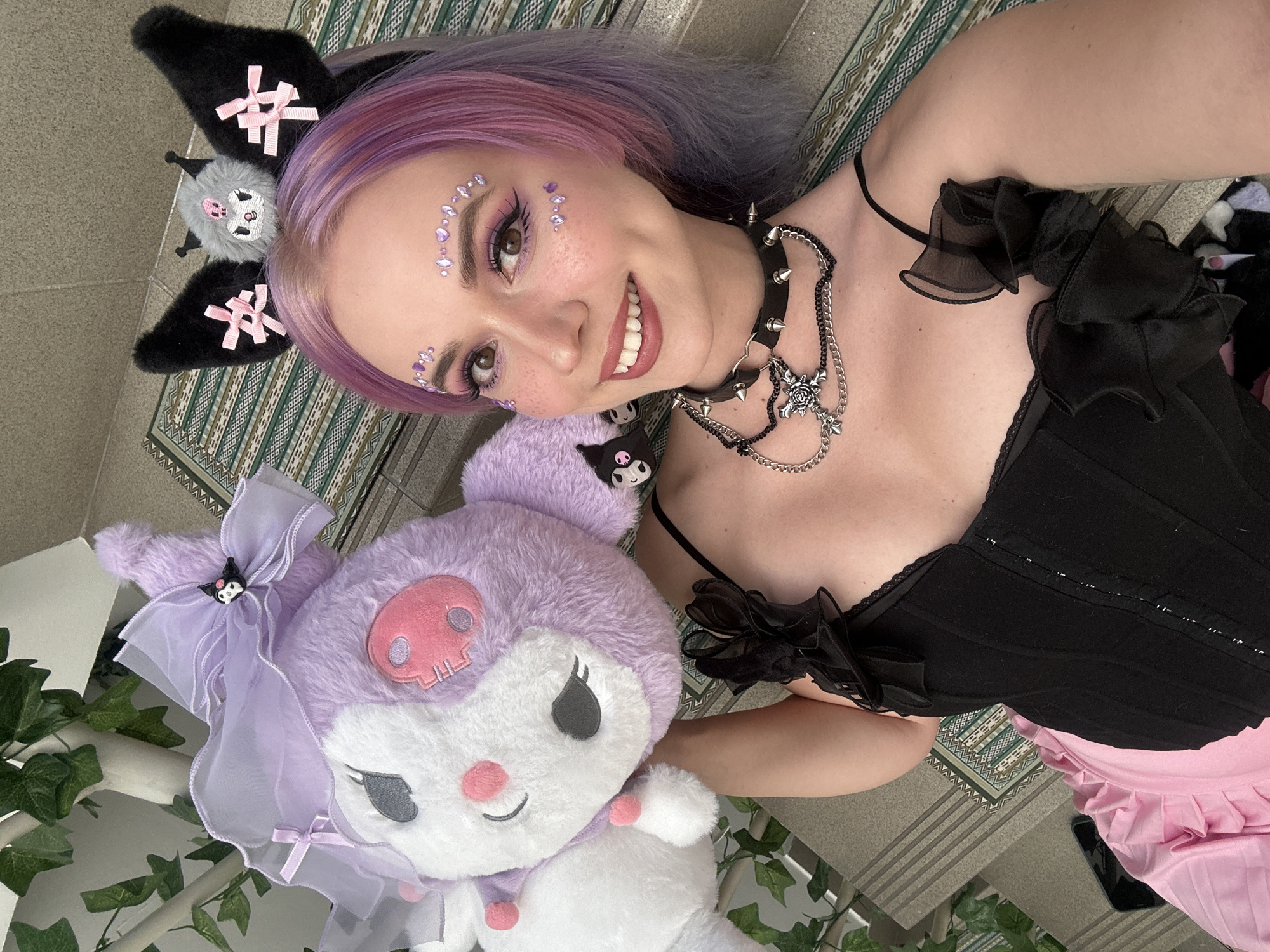 A young woman with dyed purple hair and decorative face gems is smiling and lying on a patterned surface, possibly a carpet or mat. She is wearing a black and pink outfit with a spiked choker necklace and a Kuroomi headband. She is holding a large Kuroomi plush toy, which features a lavender bow and a matching skull emblem. The background includes plants. The location of the image is unclear and unlocatable.