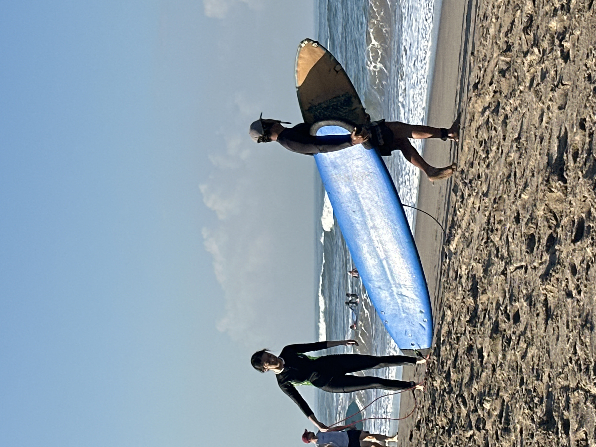 Two individuals are on a sandy beach, preparing to surf. One person, wearing a black wetsuit with green accents and a helmet, stands next to a large blue surfboard. They are holding the surfboard and appear to be looking towards the water. Another person, also in a wetsuit, is visible in the background, positioned further down the beach. They are standing next to a different surfboard, which is mostly out of frame. The ocean is in the background, with waves breaking on the shore. The sky is clear and blue, suggesting a sunny day. The sand is a light brown color and appears textured. The overall scene depicts a surfing lesson or a casual surfing session. The time of day is likely daytime, given the bright sky. The location is North Kuta, Indonesia.
