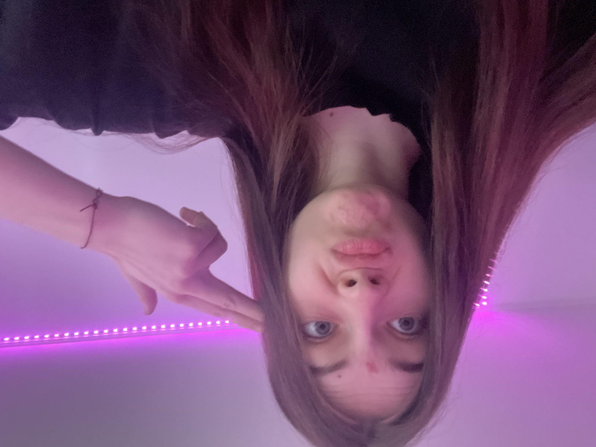 The media file features a young individual, likely female, with long brown hair, captured in a close-up, upward-angled shot. The person's face is central, with wide, focused eyes and a slightly pursed, playful expression. Their right arm is raised, with the hand forming a "finger gun" gesture pointed towards their temple. A thin, dark bracelet is visible on their wrist. They are wearing a dark top, possibly black.
The background is a uniform light purple or magenta color, primarily illuminated by a horizontal strip of bright LED lights running across the upper left portion of the image. This artificial lighting creates a vibrant, almost neon ambiance. The overall scene suggests an indoor setting, likely a room with decorative lighting. The person appears to be posing for a selfie, conveying a playful and casual mood. This scene takes place in Saint Petersburg, Russia. No visible text is present in the image.