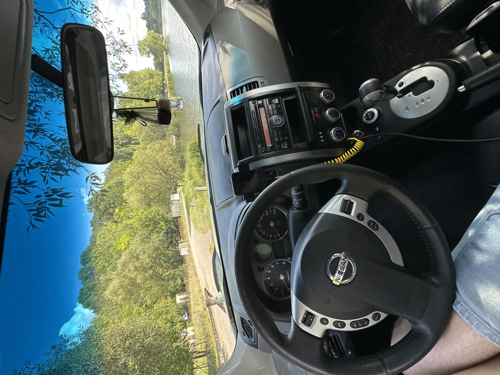 The image shows the interior of a Nissan car parked on a road with a lake and trees in the background. The steering wheel and dashboard are visible, as well as the car's audio system and gear shift. The view through the windshield reveals a sunny day with a blue sky and some clouds. The surrounding area appears to be a natural landscape, possibly a park or a rural area. There is no direct evidence of the city of Makiv, Ukraine, within the image itself.