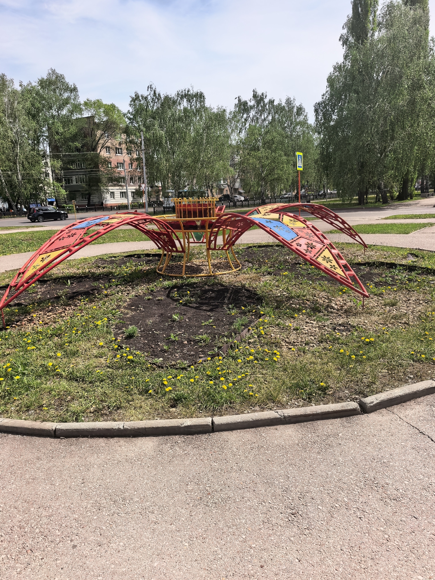 A ground-level, medium shot depicts an outdoor playground structure in Sterlitamak, Russia, on a sunny day. The playground equipment is a circular frame with multiple radiating, arching red supports, some adorned with yellow and blue triangular panels. The central structure is yellow with a cage-like top. The entire piece is situated in a circular, unkempt grassy patch speckled with yellow dandelions, bordered by a grey concrete curb. Beyond this, a faded pink asphalt path is visible. In the background are various trees, a street with vehicular traffic, a pedestrian crossing sign, and apartment buildings. The lighting is bright, casting shadows and highlighting the colors of the playground equipment.