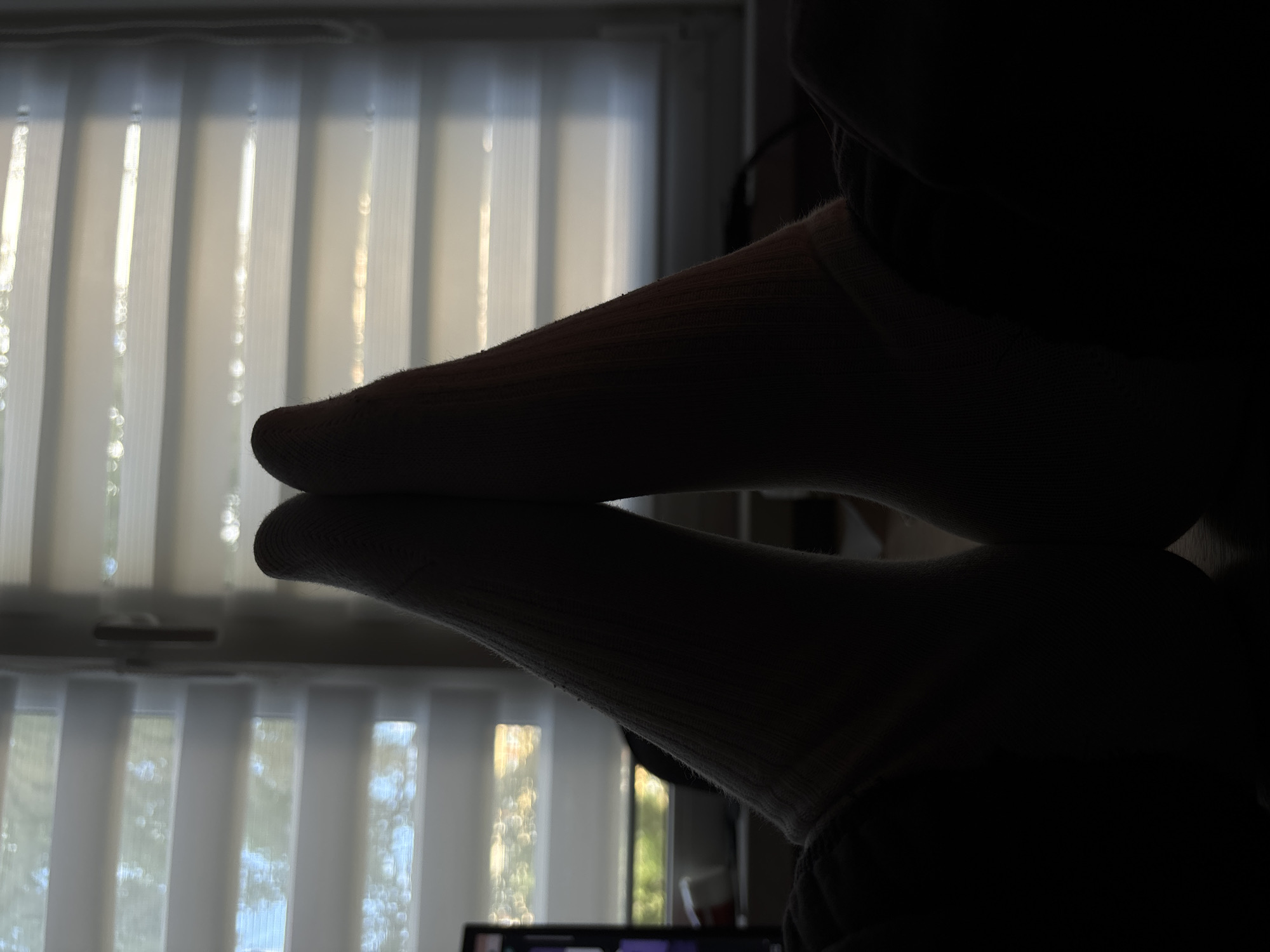 The image shows a close-up, silhouetted view of two feet wearing white socks. The feet are positioned close to each other, with one slightly above the other, creating a mirror-like effect, suggesting they are resting on a reflective surface. The background is out of focus and appears to be a window covered with vertical blinds. Light streams through the blinds, creating vertical lines of illumination. The overall mood is calm and still. The time of day is likely daytime due to the natural light coming through the window. No text is visible in the image. The setting appears to be indoors, possibly a home or office, given the presence of blinds and a hint of a computer screen in the lower portion of the image. The scene suggests a moment of rest or relaxation.