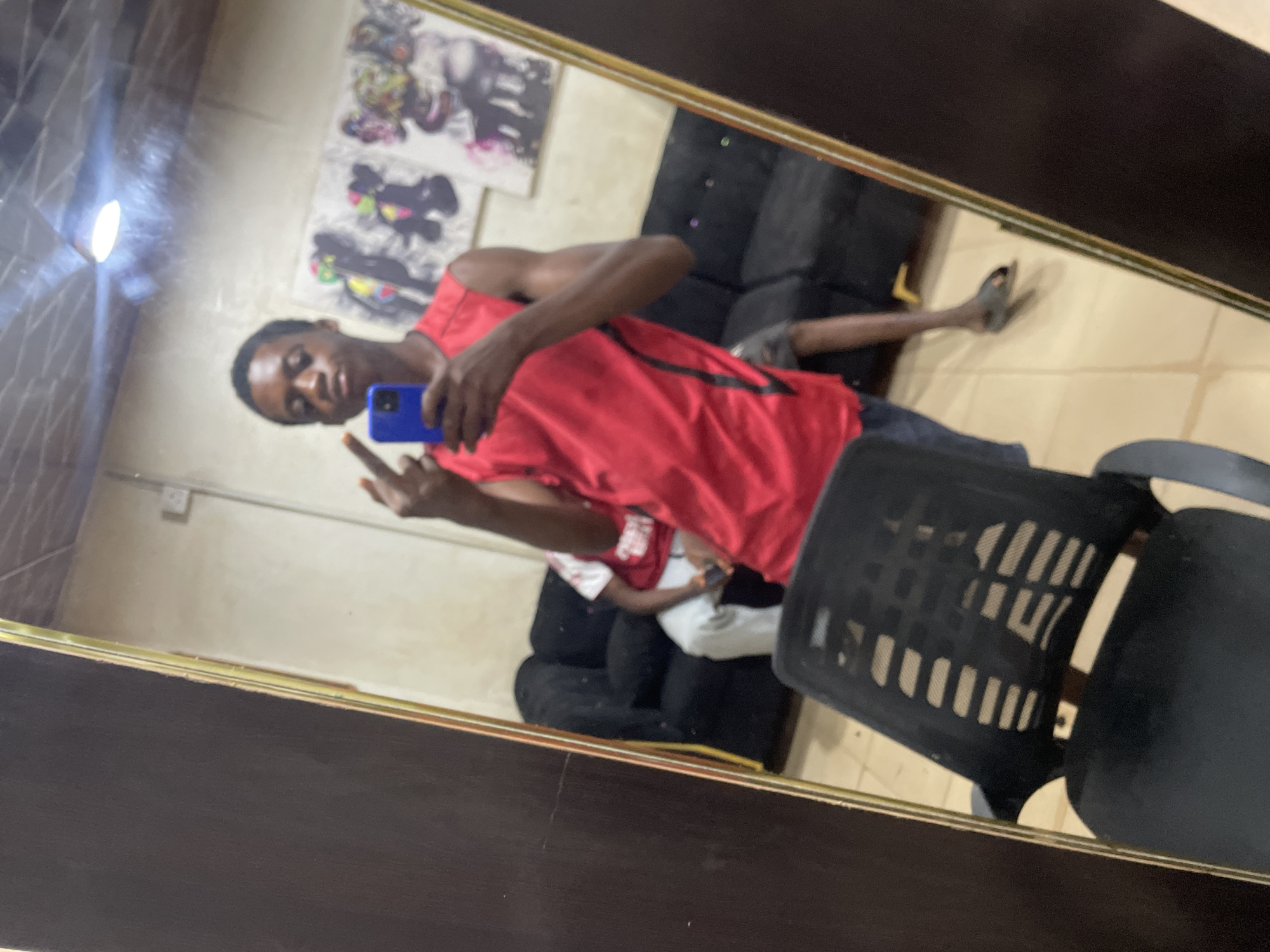A man is taking a selfie in a mirror. He is wearing a red t-shirt. He holds a blue phone in his right hand and is pointing his index finger. In the background, there is a black couch and a person sitting on it. There are posters on the wall and a black chair to the right of the mirror. The image was taken indoors. Location: Onitsha, Nigeria.