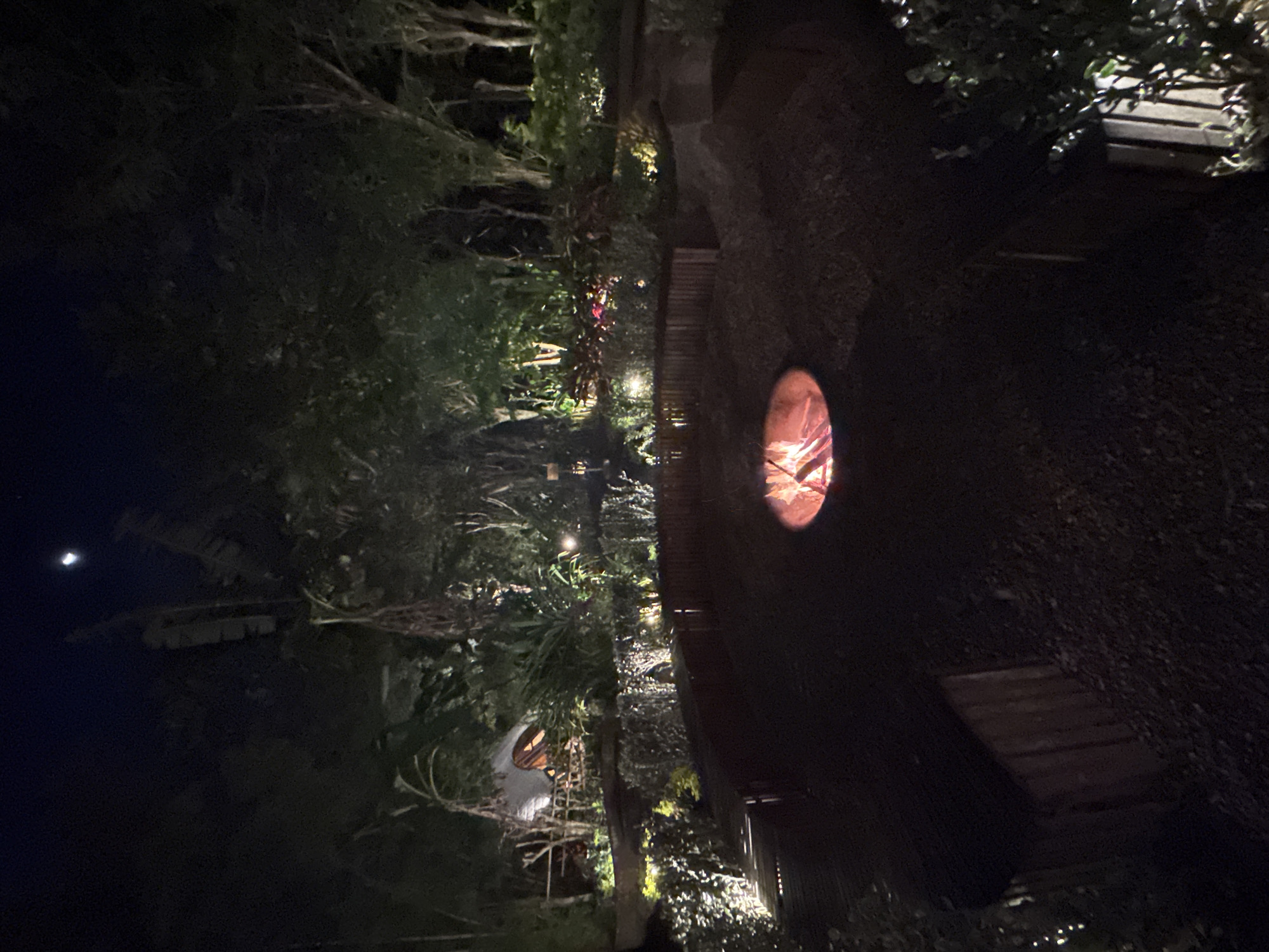 At night in Kuta Selatan, Indonesia, the image captures an outdoor space with a fire pit as a central element. The fire pit emits an orange glow, suggesting it is actively burning. The surrounding environment consists of lush vegetation, trees, and shrubs. A wooden pathway or structure is visible, possibly for seating or walking. There's low-level ambient lighting throughout the area, and the night sky with a sliver of the moon.