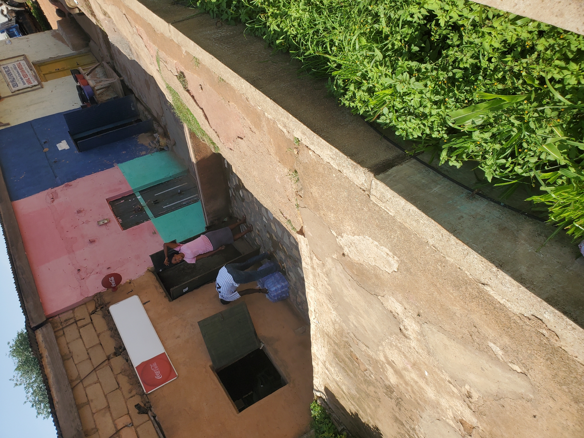 This is an outdoor scene in Bukuru, Nigeria, featuring a building with a facade of varying colors, including pink, blue, and teal. The building has multiple windows and doors, some of which are open. A man wearing a striped shirt and dark pants is seen carrying a large plastic water container. A woman in a pink shirt and gray skirt stands nearby. In front of the building, there is a paved area with some scattered debris. To the right of the building, a concrete channel is visible, filled with lush green vegetation. A Coca-Cola sign is displayed on the building. The lighting suggests it is daytime, with clear skies.