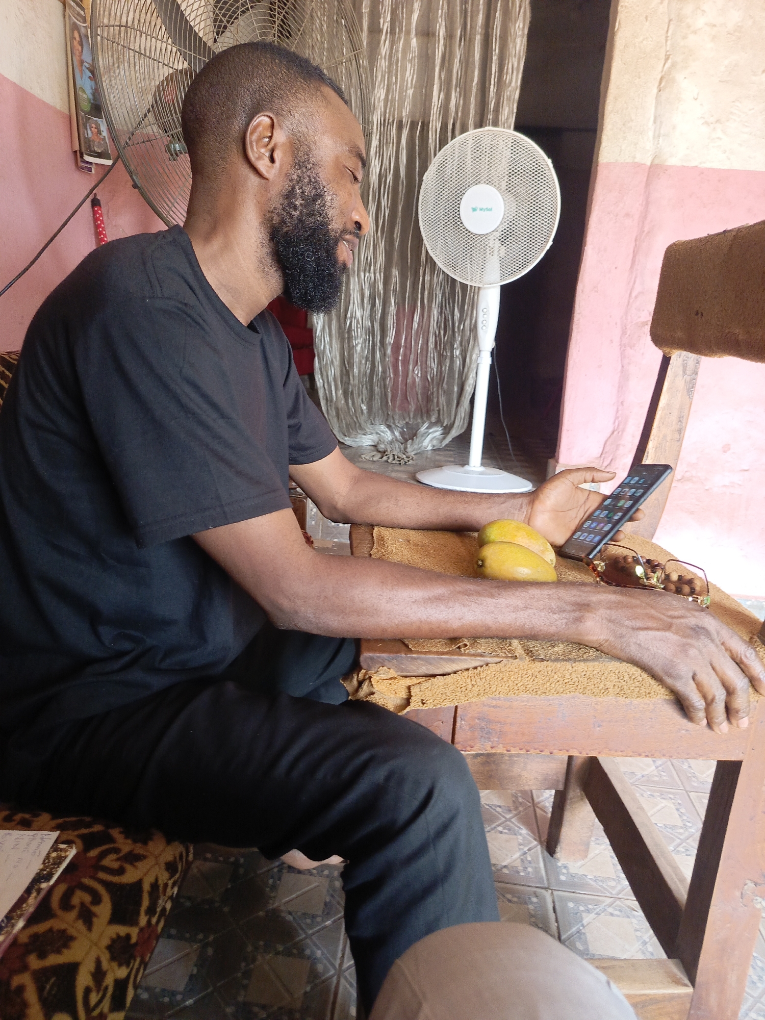 A man with a beard, wearing a black shirt and pants, sits on a wooden chair inside a building in Doma, Nigeria. He is looking at a smartphone in his right hand. Two mangoes and a pair of eyeglasses are on the chair beside his hands. A floor fan is behind him, and a larger oscillating fan is visible in the background. The walls are painted pink and off-white.