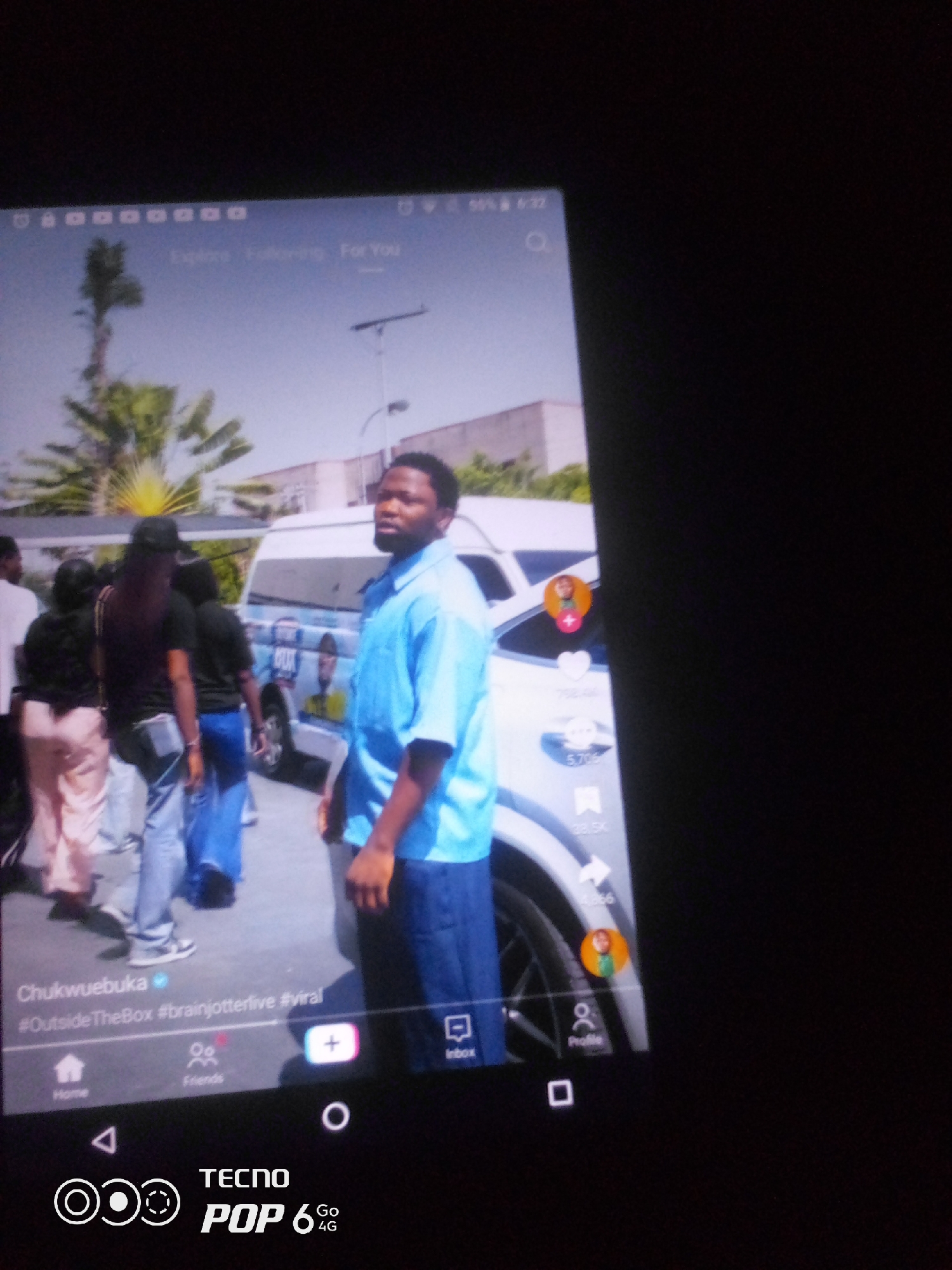 The image shows a screenshot of a social media application on a TECNO POP 6 4G phone. The screen displays a video featuring a young Black man in a blue shirt and jeans, standing in front of a white van. He is looking towards the right side of the frame. Behind him, several other individuals are visible, some appearing to be female, dressed in casual attire. One person is wearing a pink outfit, another in a black top and blue jeans, and another in a black top and a black headscarf. The environment appears to be outdoors, with palm trees and a building visible in the background, suggesting a warm climate. The interface elements of the social media app, such as icons for "Home," "Friends," "Inbox," and "Profile," are displayed at the bottom of the screen. The video's description includes the text "Chukwuebuka #OutsideTheBox #brainjotterlive #viral." The phone's status bar shows "50% 6:22." There is no specific information about the city of Howayi, Nigeria within the provided image.