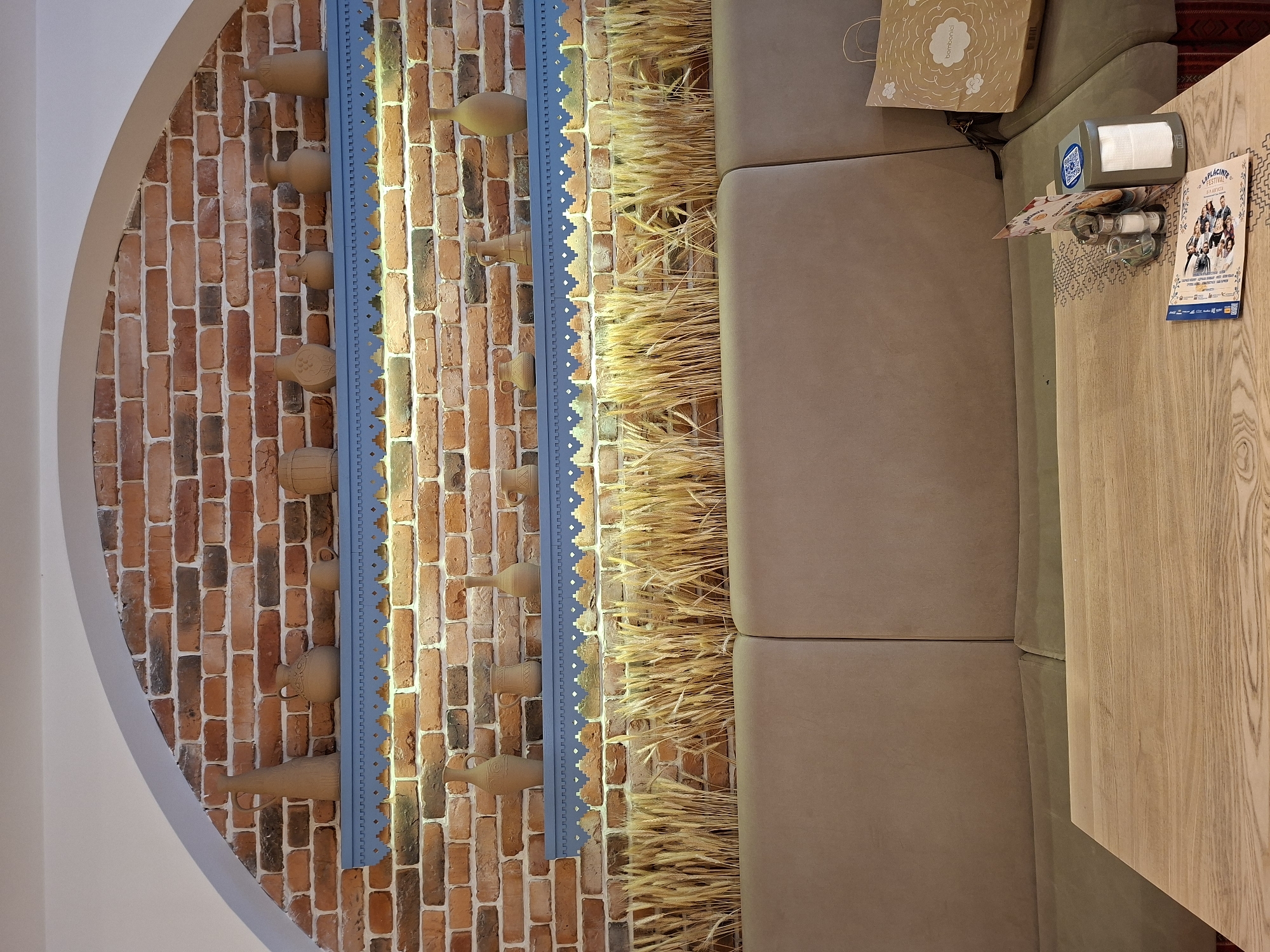 The image depicts an interior scene of a restaurant with a rustic and traditional ambiance. The background is a brick wall, with two blue decorative shelves mounted on it. The shelves hold various ceramic vases and jugs of different sizes and shapes. Below the shelves, there is a decorative element of dried wheat stalks. In the foreground, there is a cushioned seating area, likely a booth, made of a beige fabric. A wooden table is positioned in front of the seating. On the table, there are several items, including napkins, salt and pepper shakers, a menu with images of people, and a small holder with a QR code. To the right, a paper shopping bag with a cloud design and "bombonici" branding is placed on the seating. The lighting suggests an indoor setting, possibly a restaurant in Chișinău, Moldova, given the "bombonici" branding which is a popular confectionery brand in Moldova and commonly found in their stores and cafes.