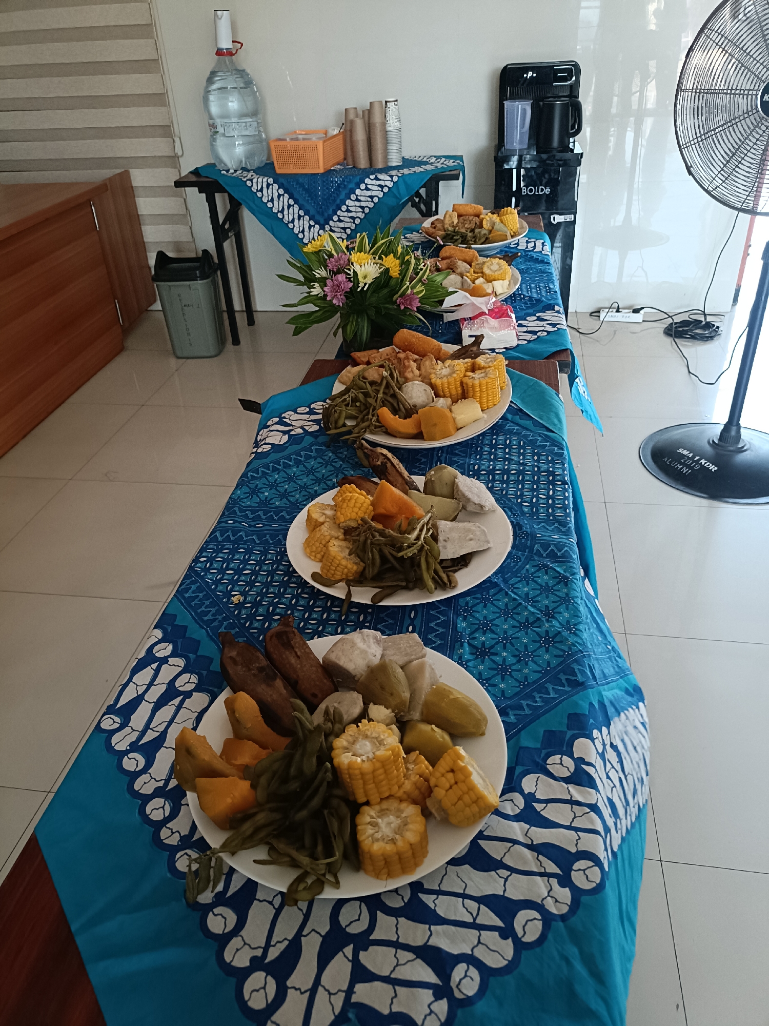 The image depicts a food spread arranged on a table draped with a blue batik cloth. The table is set up in an indoor environment, likely a public or communal space, possibly a school or office, given the presence of a water dispenser and a trash can. The food consists of various items including corn on the cob, green beans, and what appears to be steamed taro or sweet potato, presented on white plates. There are also some fried items, possibly spring rolls or similar snacks, visible on the uppermost plate. A floral arrangement adds a decorative touch. In the background, a large industrial fan is positioned on the right side of the frame, and a water cooler is visible. The floor is tiled with a light-colored pattern. The overall scene suggests a catered event or a communal meal. While the specific location in Kediri City, Indonesia, cannot be determined from the image alone, the presence of Indonesian batik and typical Indonesian food items suggests a domestic setting.