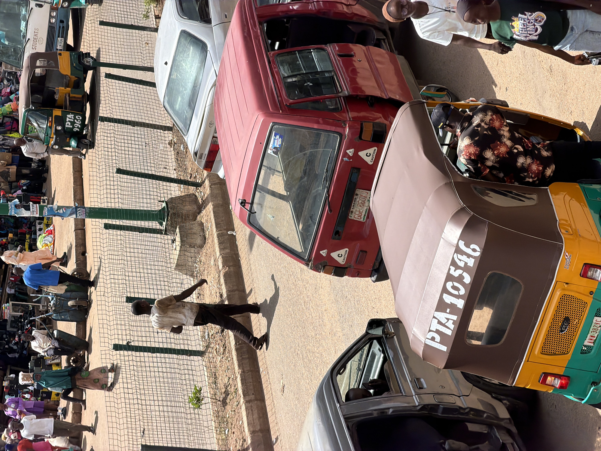 The image captures a street scene in Bukuru, Nigeria, a busy commercial area. The focus is on several vehicles and individuals. In the foreground, a yellow and green auto-rickshaw, identified with the license plate "AMV-382VC", is parked with its door open. A person wearing a black floral shirt and a baseball cap is entering the auto-rickshaw. To the right, a young woman in a white t-shirt and glasses and another in a green t-shirt are standing.
Behind the auto-rickshaw, a red minivan with an open door is also parked. On its rear, a "CAUTION" sticker is visible. To the left of the minivan, a silver sedan is parked, and a person can be seen through its window. Further back, a man in a white shirt and dark pants walks on the pavement next to a wire fence.
In the background, a larger bus with "STUDENTS" written on its side is parked. Another auto-rickshaw, predominantly green and yellow with "PTA 9960" on its side, is seen further down the road. The street appears to be paved, and there are market stalls visible in the distance, suggesting a bustling environment. The overall scene depicts a typical day in a Nigerian city, with various modes of transport and people engaged in daily activities.