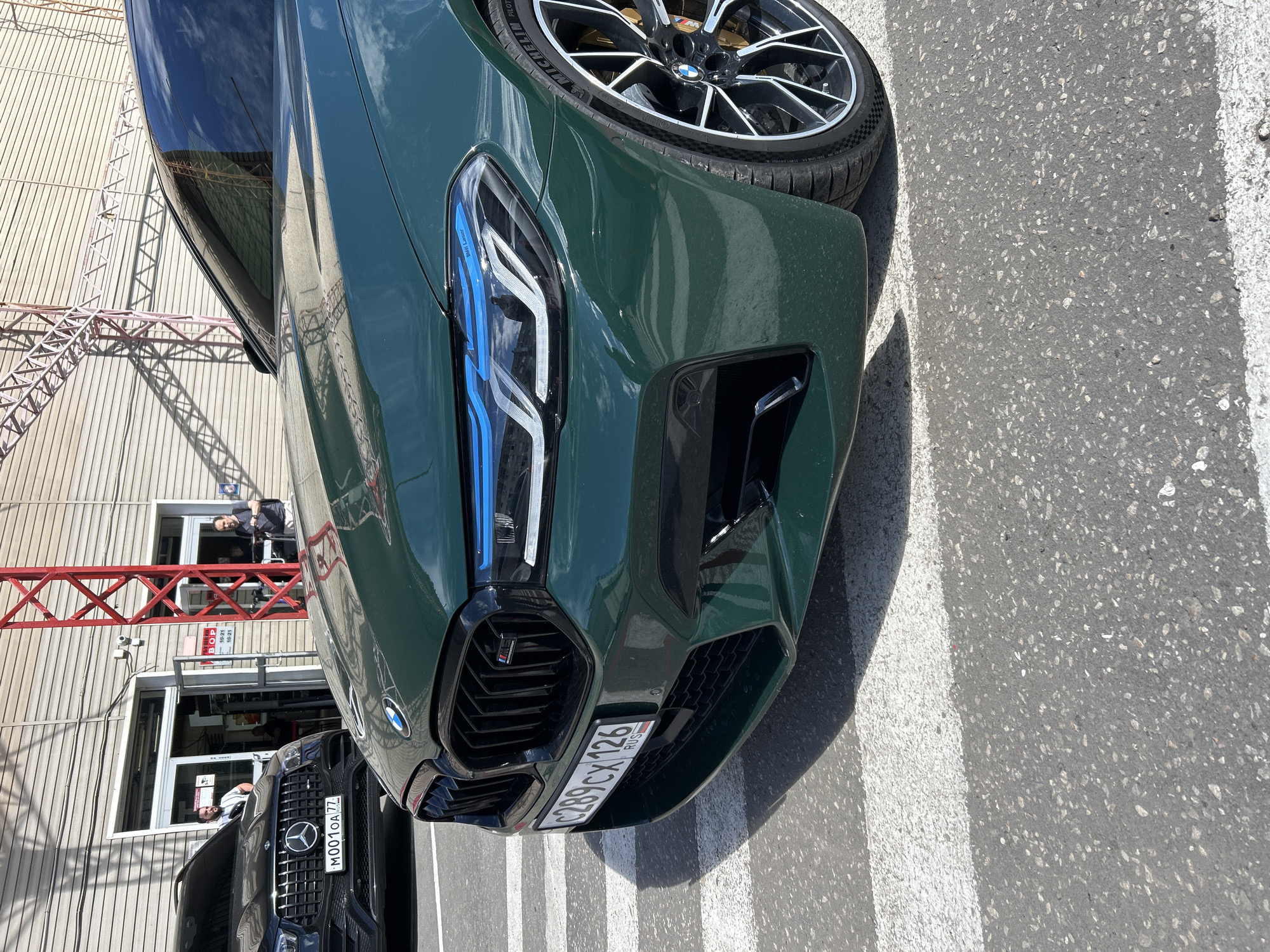 The image shows the front of a dark green BMW M car parked on asphalt, with white lines visible on the ground, suggesting a parking lot or road. The car is facing slightly to the right, with its headlights, grille, and part of the front wheel visible. The headlights have a distinctive LED design with blue accents. A license plate with Russian characters is affixed to the front bumper. The BMW logo is visible on the wheel.
In the background, there's a building with a corrugated metal exterior. A red metal structure, possibly a scaffolding or part of an industrial building, is attached to the side of the building. Two people are visible through a window of the building, one standing and looking out, and another seated. Another car, a black Mercedes-Benz, is parked in front of the building, with its license plate partially visible. The time of day appears to be daytime, with clear shadows indicating sunlight. The weather seems to be sunny. The scene is set in Moscow, Russia, as indicated by the Russian license plates.