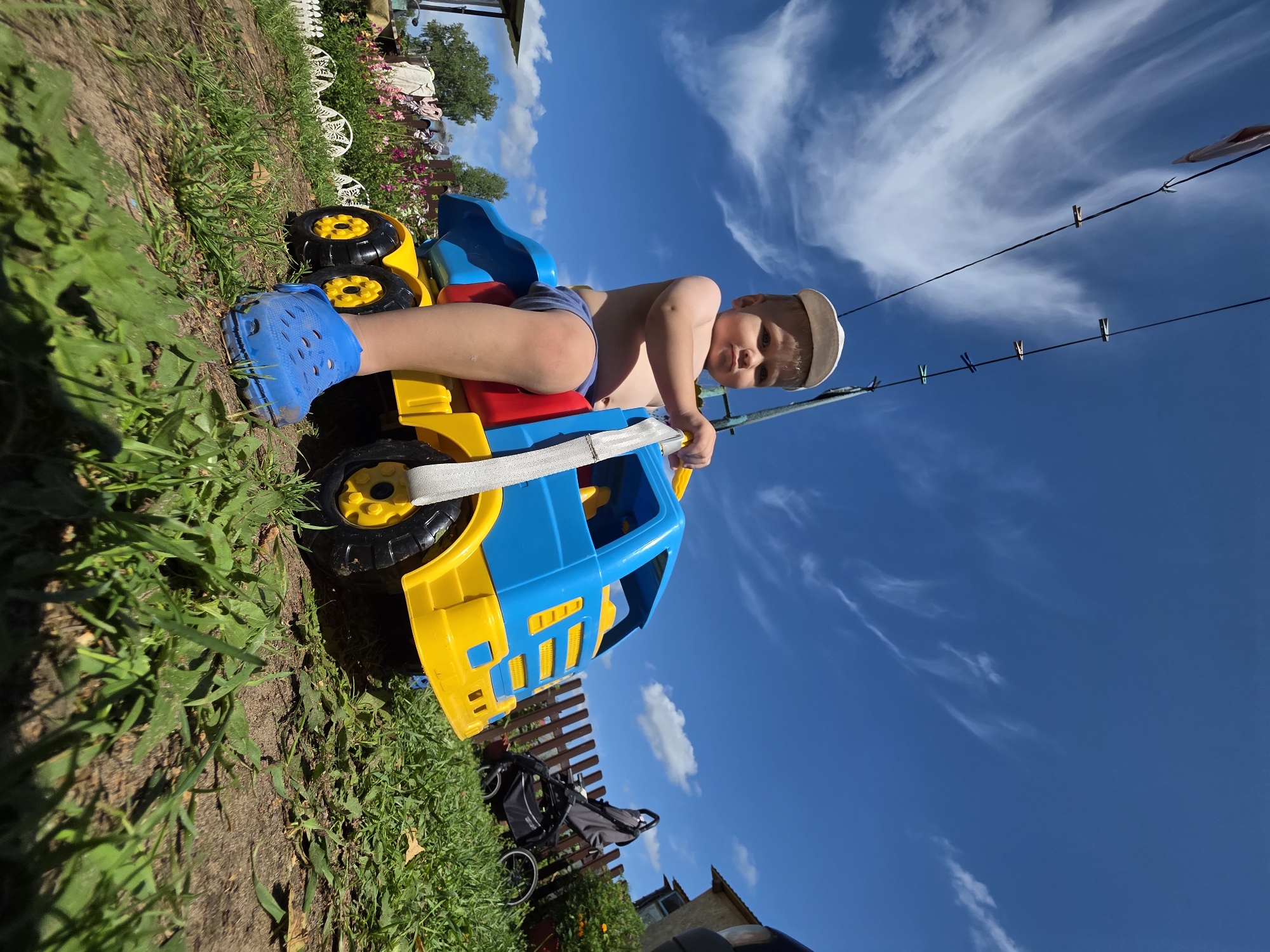 A young child wearing a white cap and blue Crocs sits on a blue and yellow toy dump truck outdoors in a grassy yard. The child is shirtless and wearing blue shorts. The truck has large black and yellow wheels. Behind the child, a brown wooden fence separates the yard from a garden with pink and purple flowers. A gray baby stroller is parked next to the fence. The sky is bright blue with wispy white clouds. Several clothespins hang on a wire in the upper left portion of the image. The scene appears to be in a rural or suburban setting in Буренка, Russia, given the style of the house and garden.