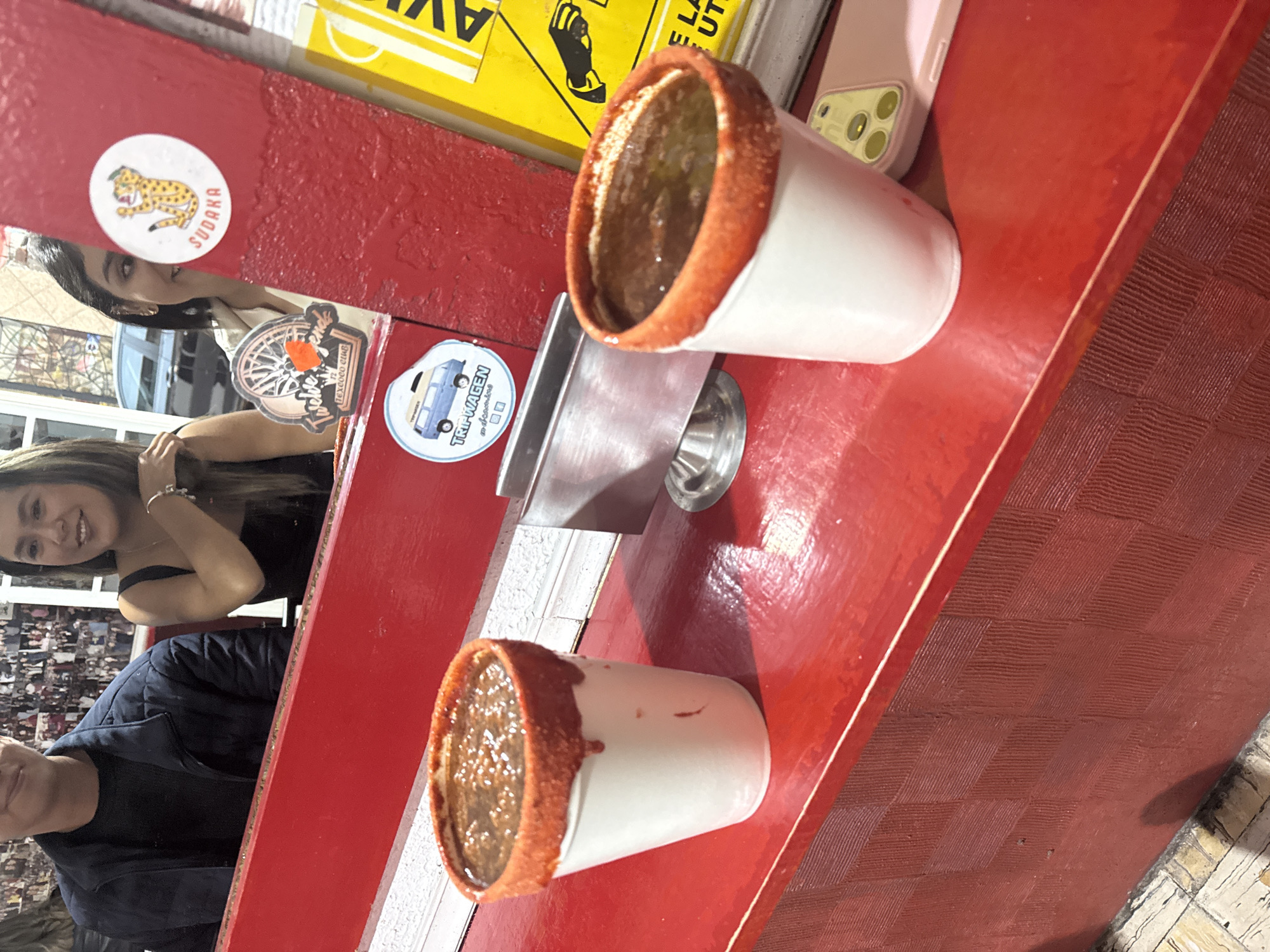 The image captures a scene at a food stall in San Andrés Cholula, Mexico. Two white styrofoam cups filled with a dark liquid, likely a michelada or similar beverage, are placed on a red counter. The rims of the cups are coated with a red spice, possibly Tajin. Behind the cups, two people are visible: a woman with long brown hair wearing a black top and a man wearing a black shirt under a dark jacket. Stickers are affixed to the red counter, including one with a jaguar and the name "Sudaka." A pink cellphone lies on the counter near the cups. The environment suggests a lively, possibly outdoor, food vending area.