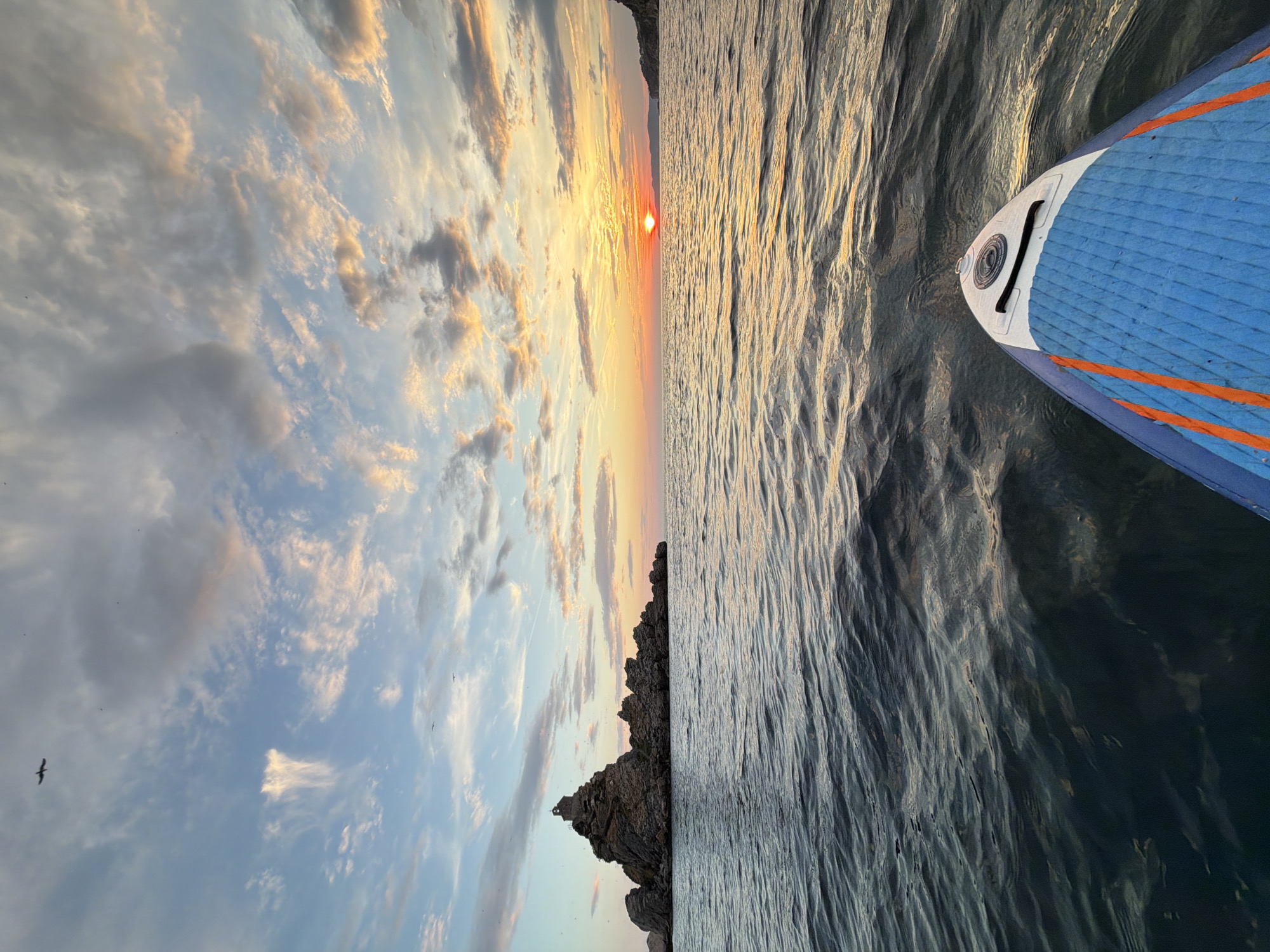 This is a photograph taken from the perspective of someone on a stand-up paddleboard, looking out towards a sunset over the sea. The image is oriented vertically.
The subjects include the front of a blue and white stand-up paddleboard with orange trim, which occupies the right side of the frame. The sea is on the right, with gentle ripples reflecting the golden light of the setting sun. On the left side of the image, a dramatic rocky outcrop juts out of the water. Above the rocks, the sky is filled with a mix of soft, fluffy clouds tinged with the warm colors of sunset - oranges, pinks, and yellows against a pale blue background. The sun itself is a bright, small orb near the horizon, casting a golden glow. A few small birds are visible in the sky.
The scene captures a peaceful moment at dusk. The light suggests it is either sunrise or sunset, with the sun low on the horizon. The weather appears calm and pleasant, with the gentle water and soft clouds. The rocky outcrop, likely a small island or part of the coastline, provides a sense of location, hinting at a coastal environment, potentially near Petrovac centar, Montenegro, as suggested by the location context. The overall mood is serene and contemplative, conveying the beauty of nature at the end of the day. There is no visible text in the image.