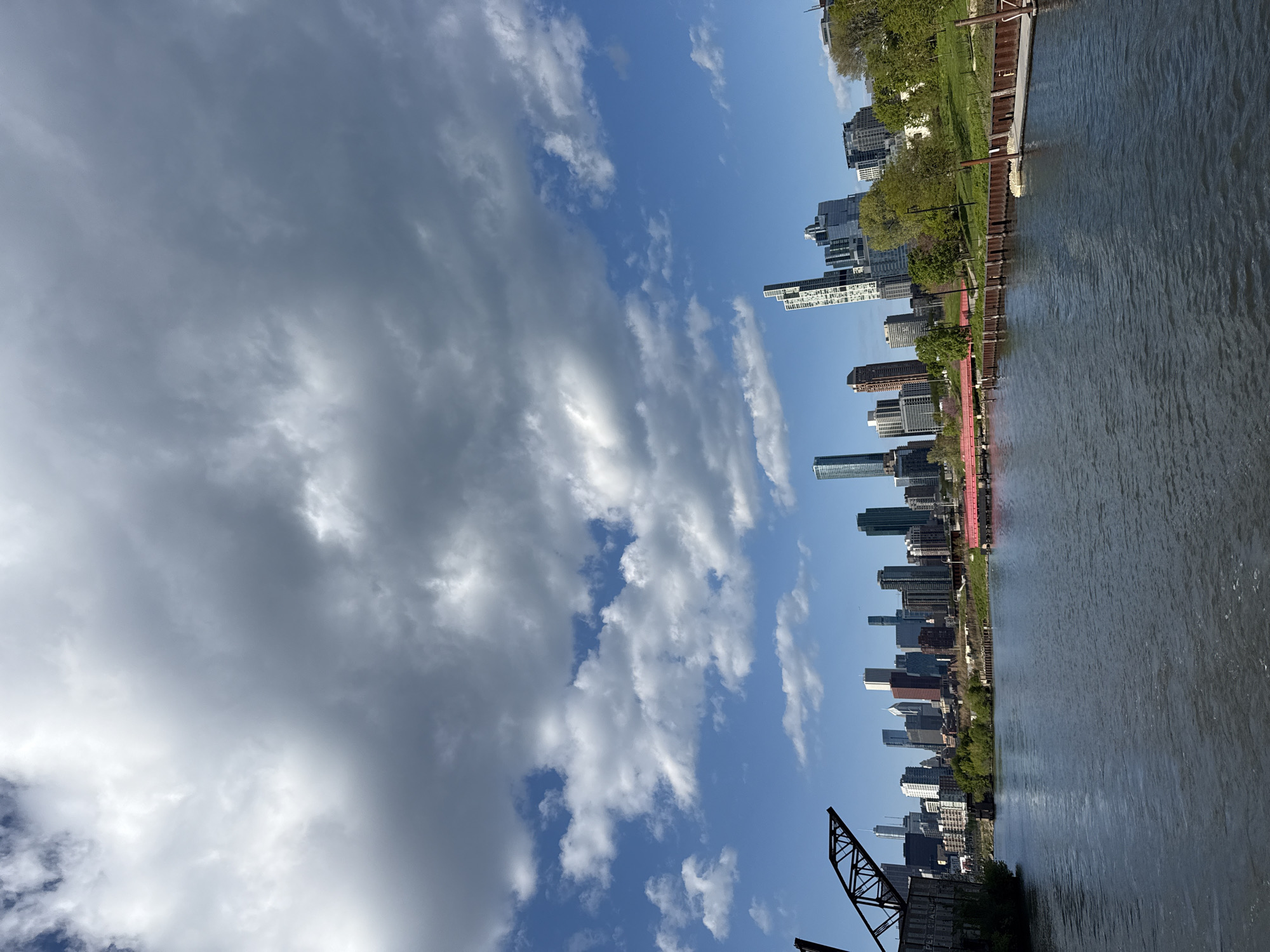 The image depicts a view of the Chicago skyline from a vantage point over the Chicago River. The photograph captures a portion of the river in the foreground, with a dark water surface reflecting some light. Along the riverbank, there is evidence of vegetation, grass and trees, and the city's infrastructure, including a red pedestrian walkway. The backdrop showcases the city's architecture, including high-rise buildings and skyscrapers, of varying heights and styles. The sky features blue patches interspersed with cumulus clouds of varying densities and formations, suggesting a partly cloudy day. At the bottom left corner, elements of a metal structure resembling an industrial crane or bridge component are visible. This view is common along the Chicago River, where industrial history meets modern urban development.