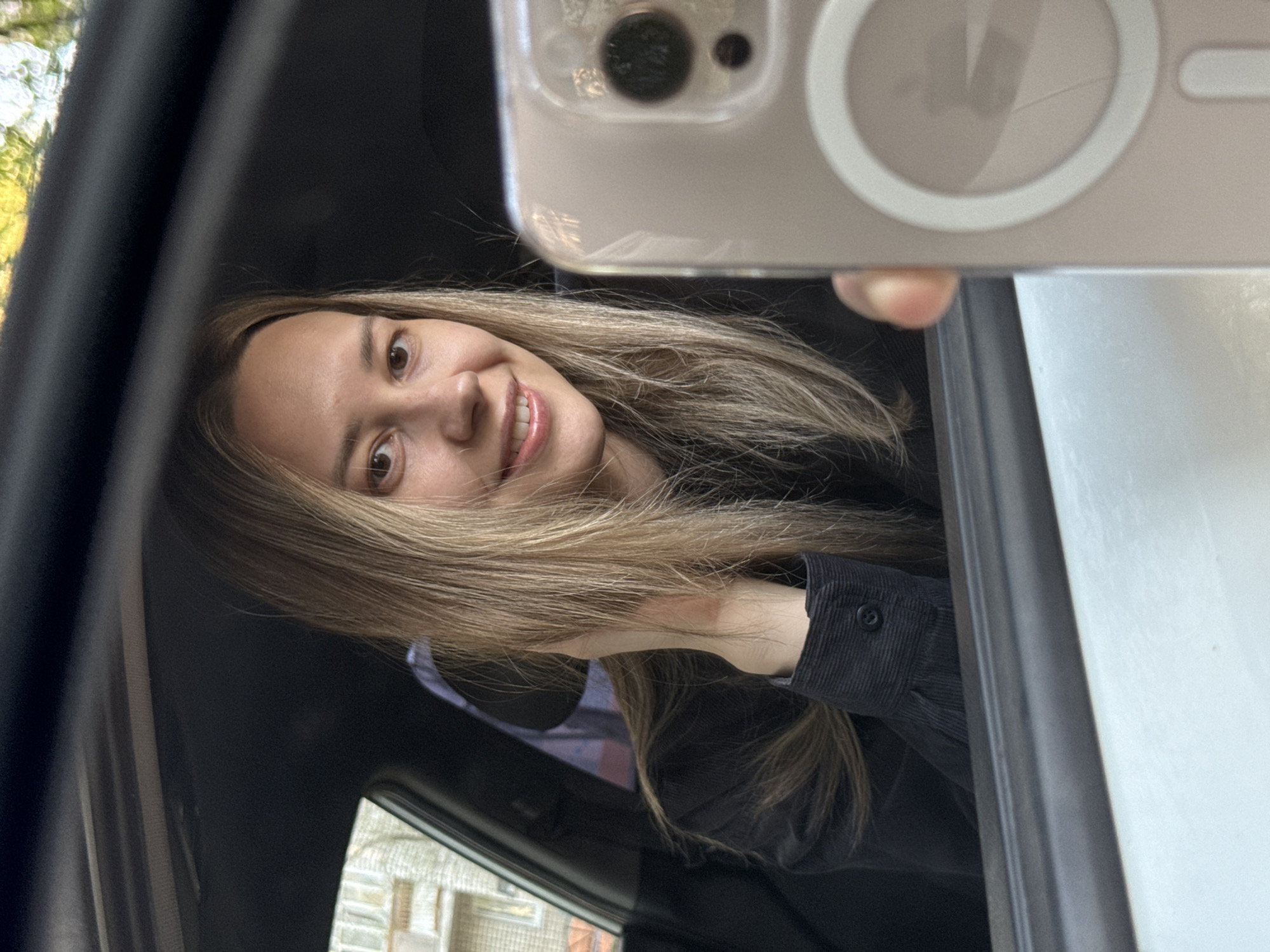 A light-skinned woman is taking a selfie with a white smartphone in a vehicle. The woman has brown hair and is smiling. She is wearing a dark-colored shirt. The background shows a blurry cityscape, which suggests that the image was taken in Chaikovsky, Russia. The car interior is dark. The focus is on the woman and her phone.
