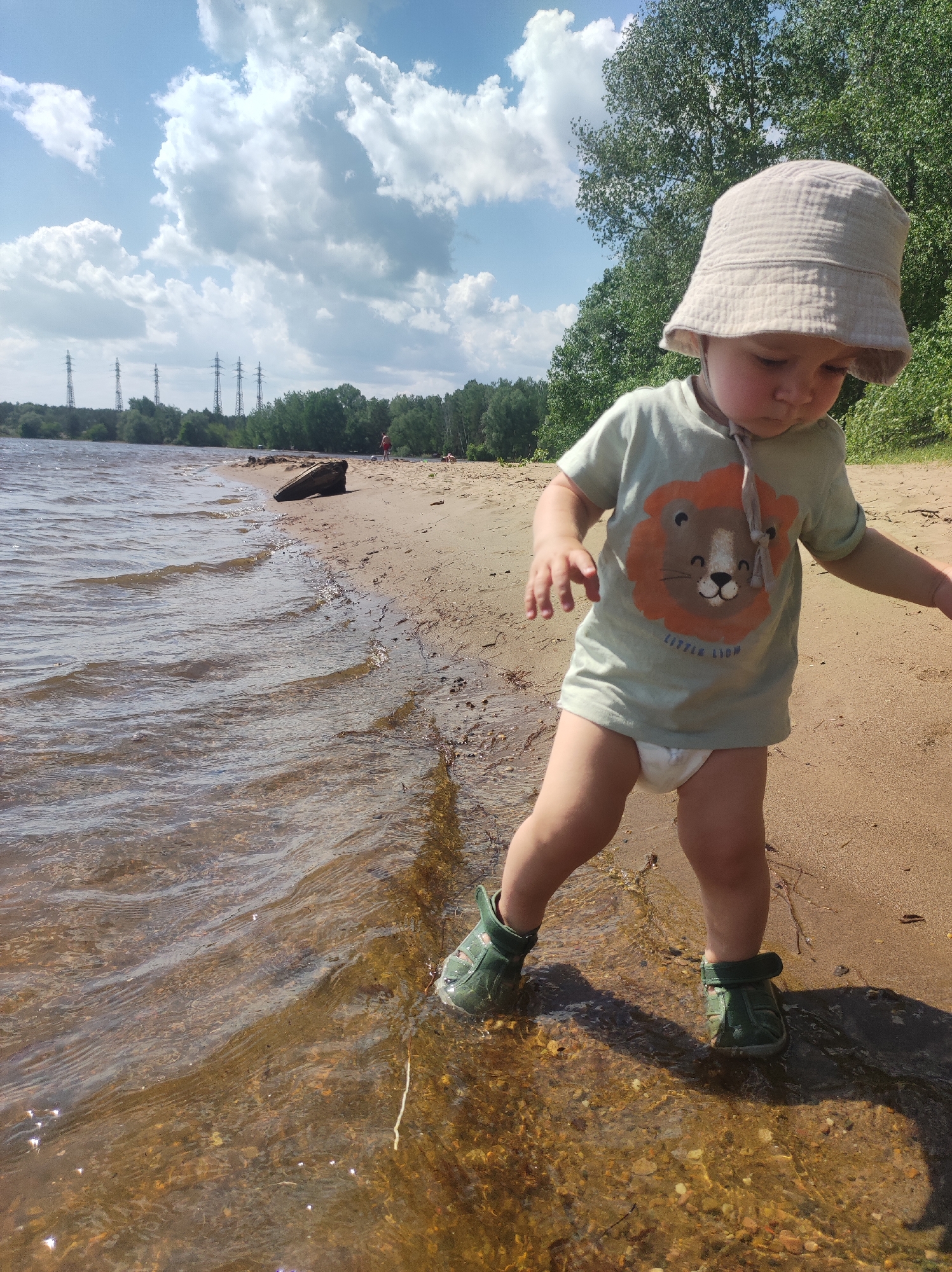 A young child, likely a toddler, stands on a sandy beach at the edge of a body of water, possibly a lake or river. The child wears a light-colored bucket hat, a short-sleeved shirt with a lion graphic, a diaper, and green shoes. The child is looking down and appears to be playing in the water or sand. The water is clear enough to see the stones at the bottom.
In the background, the scene includes a sandy beach, trees, and transmission towers visible in the distance. The location is Noviy, Russia. The sky is partly cloudy.