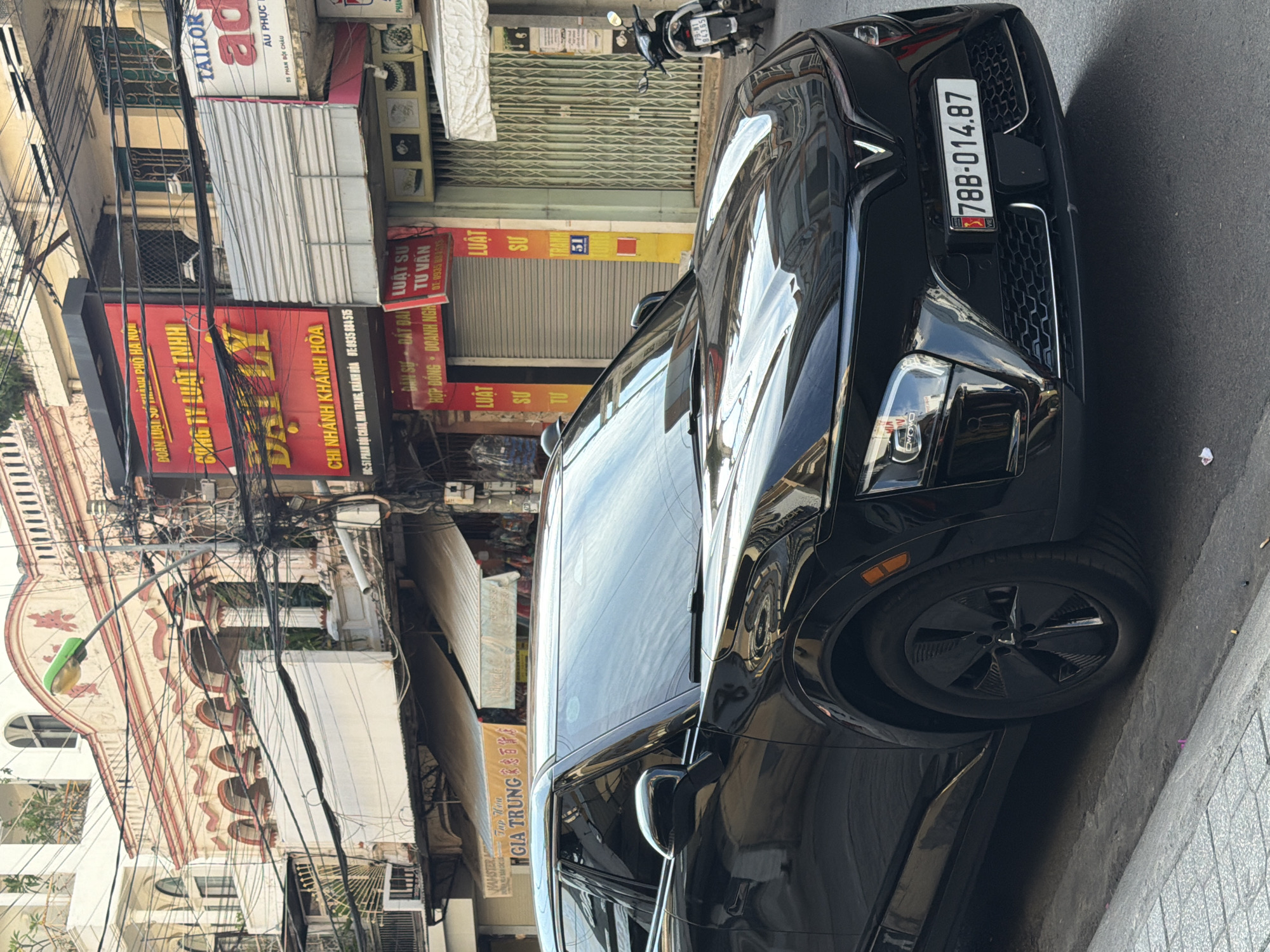 A black VinFast VF8 electric SUV is parked on a city street in Nha Trang, Vietnam. The vehicle's license plate reads "78B-014.87". The car is parked on the right side of the street, partially on the sidewalk. In the background, there are multi-story buildings with commercial establishments on the ground floor. Signs visible include "TAILOR", "GIA TRUNG", and various Vietnamese text, suggesting a commercial area. A motorcycle is parked on the street to the right of the SUV. The street appears to be an urban environment with overhead wires and utility poles visible. The weather seems to be clear.
