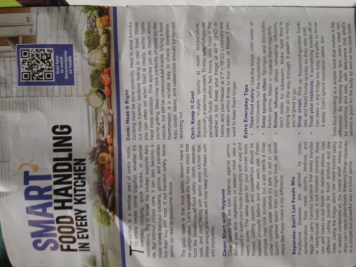This image is a photograph of a printed page, likely from a magazine or brochure, laid flat on an unidentifiable surface. The page features an article titled "SMART FOOD HANDLING IN EVERY KITCHEN" prominently displayed in large, dark blue lettering across the top left. Below the headline, a color photograph or illustration depicts an array of fresh vegetables and some prepared ingredients on a light-colored countertop, indicative of a kitchen setting. Visible items include heads of cabbage or lettuce, bell peppers, tomatoes, and other produce, some arranged in bowls. In the upper right corner of the page, a blue rectangular box contains a black and white QR code with the text "Scan here to watch documentaries on health" beneath it. The remainder of the page consists of several columns of text, segmented by headings such as "Cook: Heat It Right," "Chill: Keep It Cool," "Clean: Start With Hygiene," "Separate: Don't Let Foods Mix," and "Extra Everyday Tips," providing food safety advice. The photograph itself is slightly blurry in areas, with minor glare, suggesting it was taken under ambient lighting. No individuals, specific actions, or direct interactions are depicted within the image content. There are no visual cues or textual information within the image that indicate its location in the city of Mgbidi, Nigeria, or any other specific geographic place of origin or capture.