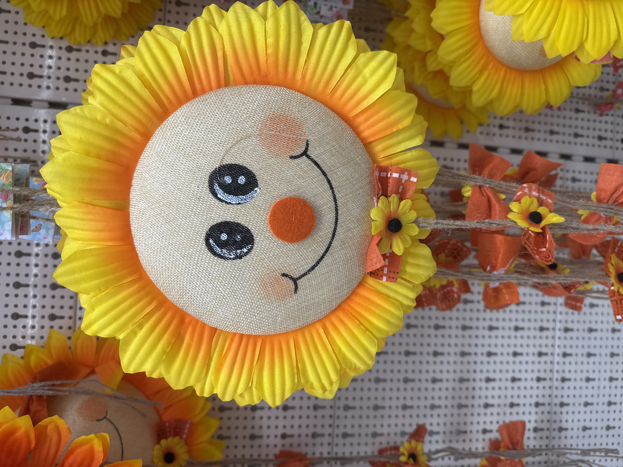 A decorative, stuffed sunflower face with a burlap center hangs from a pegboard display. The sunflower has bright yellow and orange petals, two black button eyes, a round orange nose, and a stitched smile. Two pink circles are drawn on its cheeks. A small orange bow with a miniature sunflower and white ribbon is tied at the bottom of the face. Strands of twine hang down from the bow, adorned with more orange bows and small sunflowers. The background is a white pegboard with a grid of small holes. Other similar sunflower decorations are visible to the left and right. The scene is likely in a retail store.