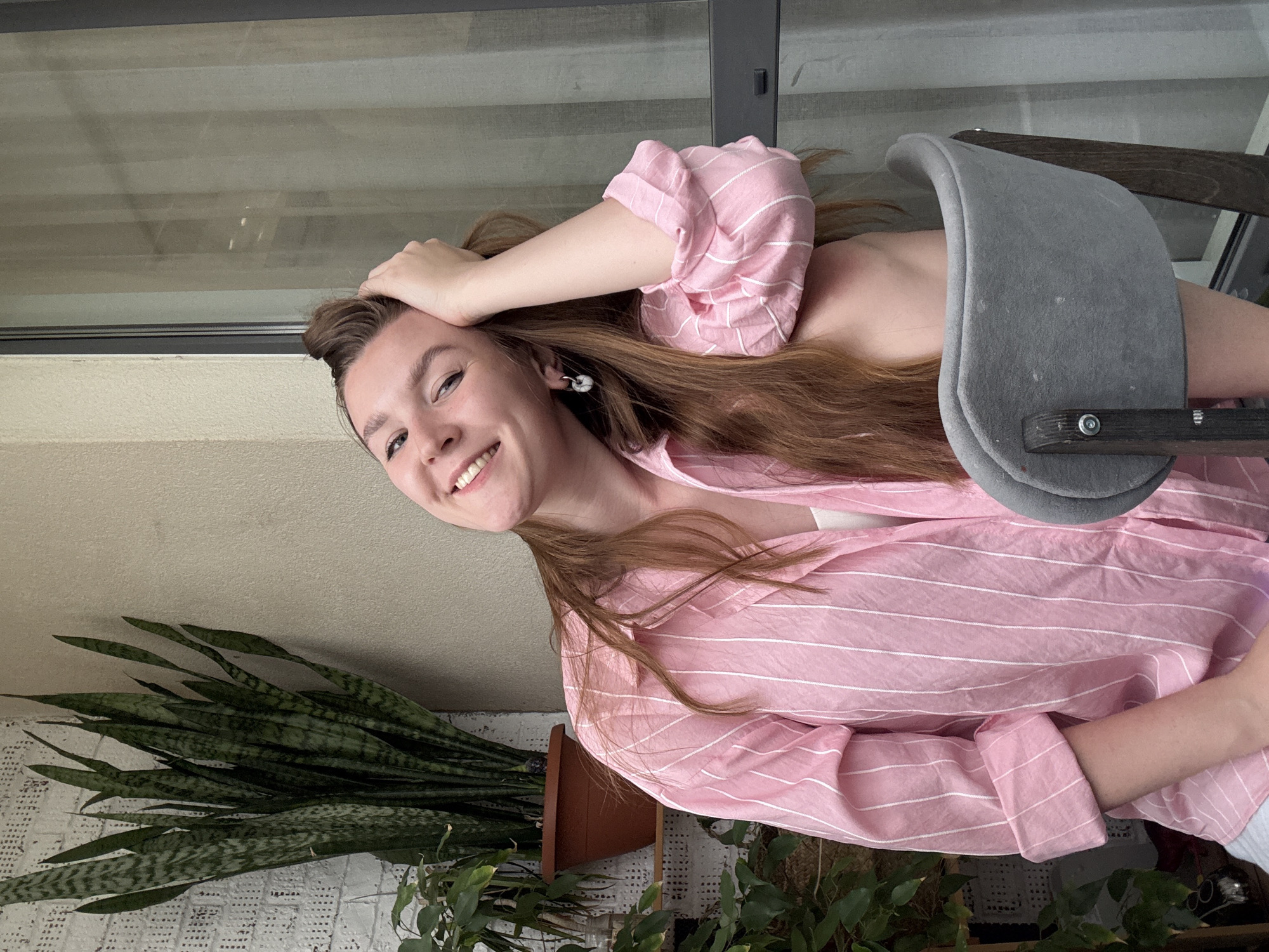 A young woman with light brown hair and fair skin is smiling at the camera. She is wearing a pink and white striped shirt. Her left hand is on her head. She is sitting in a gray chair and appears to be on a balcony, indicated by the presence of a potted snake plant and other greenery, as well as a window or glass partition in the background.
Given the information provided, pinpointing this location to Izhevsk, Russia, cannot be done based on the visual information alone.
