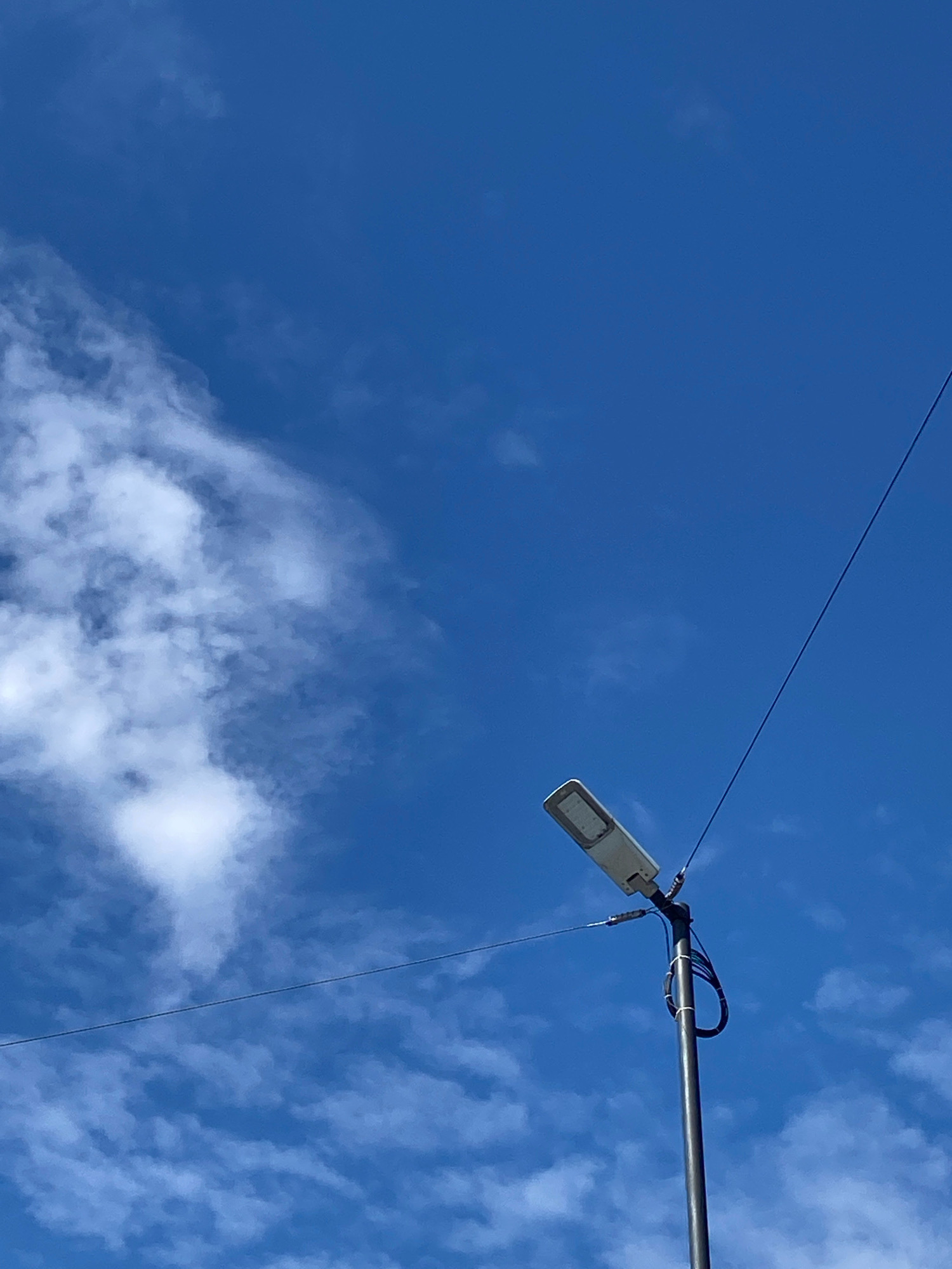 An outdoor, medium shot captures a streetlamp against a clear blue sky with scattered white clouds. The streetlamp, positioned slightly off-center to the right, is a modern, rectangular fixture mounted on a dark pole. Wires extend from the top of the pole, one running diagonally upwards and to the right across the frame. The lower portion of the pole shows a coiled black cable secured with white ties. The sky is a vibrant blue, with wispy white clouds forming on the left side of the image and smaller, more dispersed clouds further to the right. The lighting suggests daytime, likely a sunny or partly cloudy day. The location is the city of Unknown, Unknown.