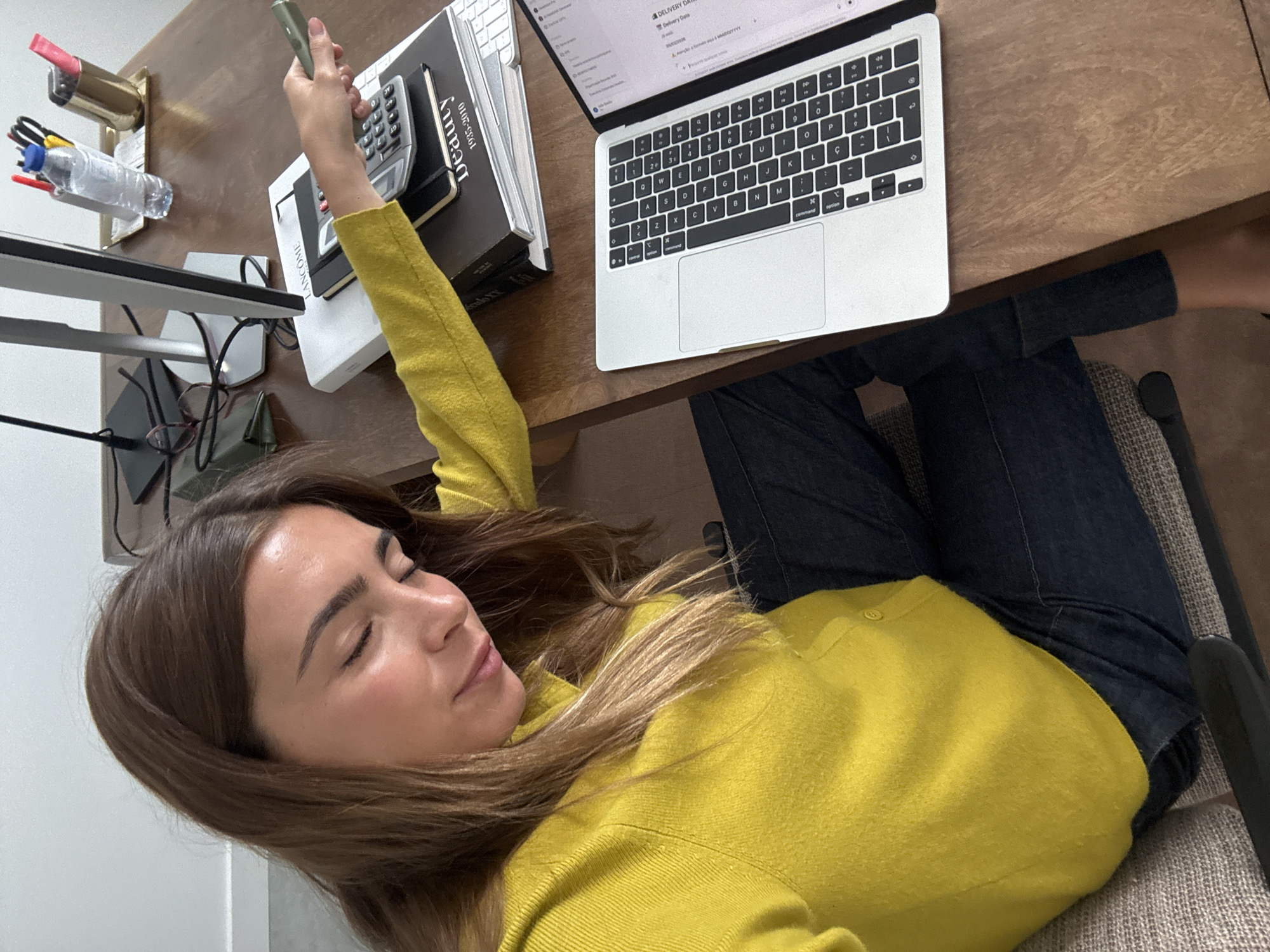 A woman in a yellow sweater is lying down at a desk. She is holding a small, grey object in her hand. On the desk is a laptop, a stack of books, a calculator, and a water bottle. The woman is wearing dark jeans and sitting in a grey chair. There is no indication that the image was taken in Torres Vedras, Portugal.