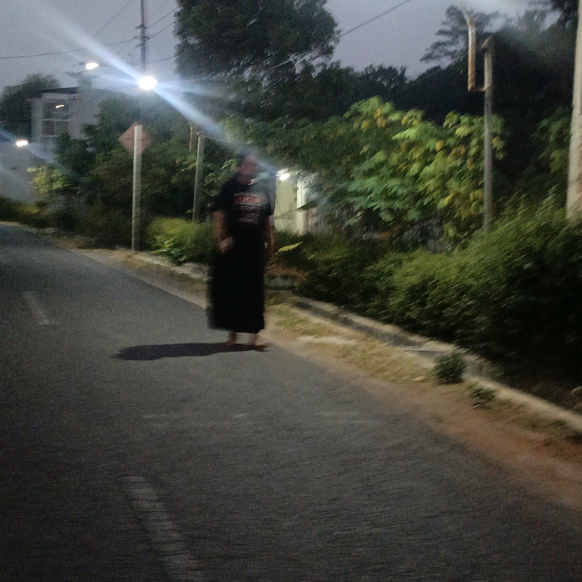 The image depicts an outdoor scene on a road at dusk or night, likely in Salatiga, Indonesia, as indicated by the presence of lush vegetation and what appears to be a residential building with architectural elements common in the region. A person, wearing a long black garment that reaches their ankles and a t-shirt with a graphic design, stands on the asphalt road. The person's face is indistinct due to motion blur and low lighting, but their posture suggests they are standing still or walking slowly. They are barefoot.
To the left of the road, a white building is visible, partially obscured by trees and vegetation. Streetlights cast bright beams, creating glare and lens flares that illuminate the scene. A traffic signpost with a diamond-shaped sign is planted on the left side of the road. The road itself has white markings, and the surrounding area is characterized by dense greenery, including bushes and trees, typical of the tropical climate. The ground on the right side of the road appears to be a dirt or gravel embankment with some dry grass and sparse vegetation. The overall impression is that of a quiet, possibly residential street at the edge of a more natural or undeveloped area, illuminated by artificial lighting in the fading light.