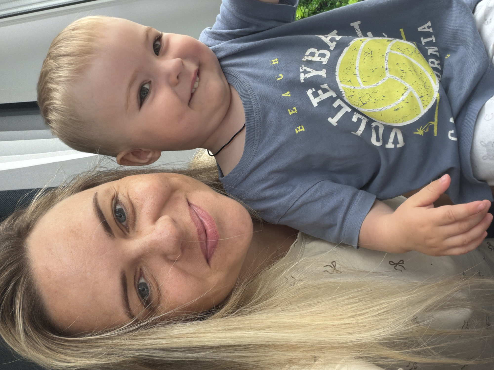 This is a medium shot selfie taken outdoors, likely on a balcony, with a woman and a young child in the foreground. The woman, on the left, has long blonde hair and is smiling at the camera. The child, on the right, is also smiling and wearing a blue t-shirt with a yellow volleyball graphic and the text "BEACH VOLLEYBALL CALIFORNIA." The background shows vertical blinds and some greenery. The image appears to be taken during daylight hours. There are no specific landmarks in the image that would indicate its location within Votkinsk, Russia.