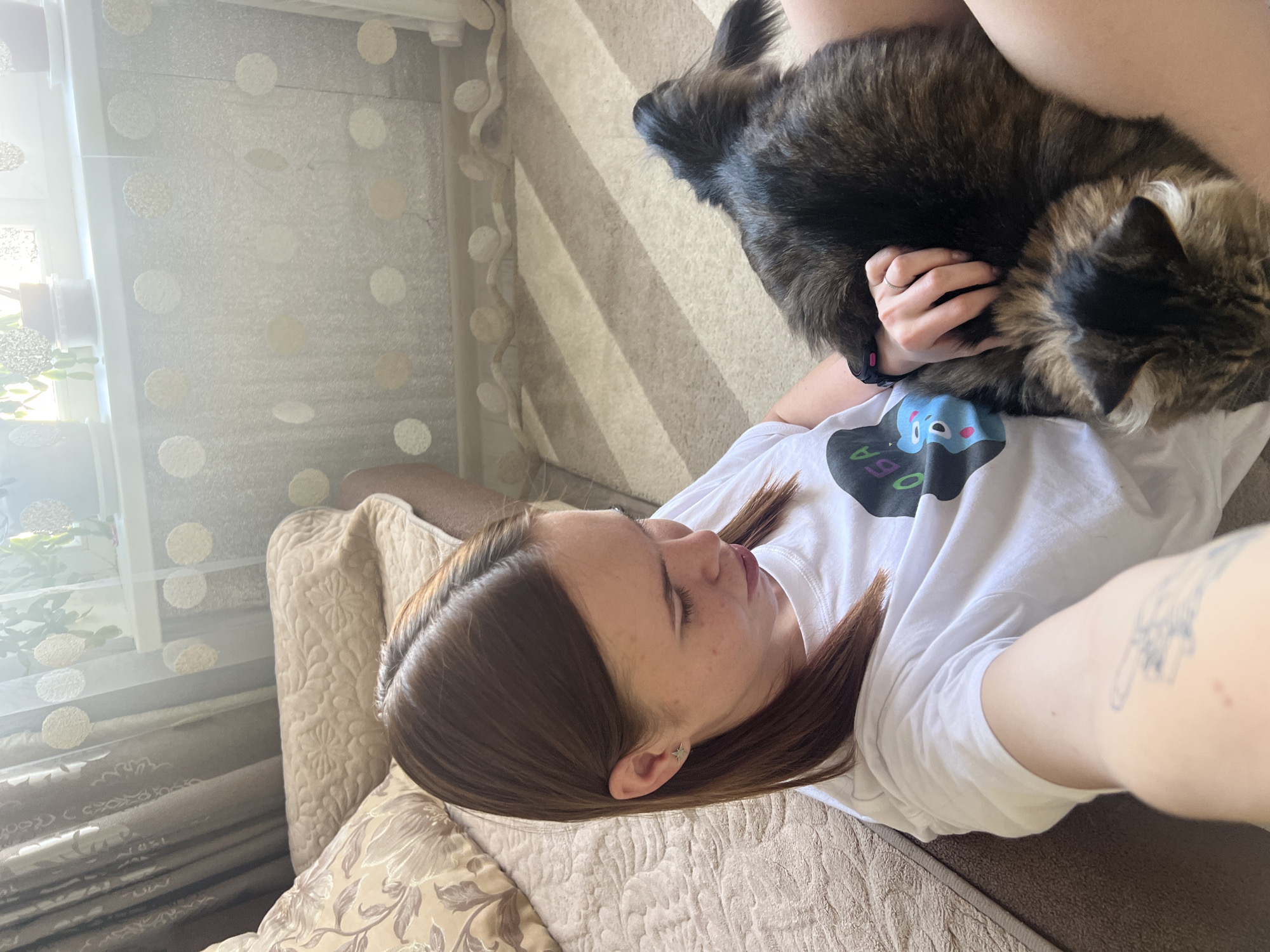 A woman lies on a beige floral couch in what appears to be an apartment. She has brown hair and is wearing a white T-shirt with a graphic design on it. A large, dark-furred cat rests on her chest and upper abdomen. A hand reaches up to hold the cat. A striped pillow is partially visible behind her head. In the background, there is a window covered with a sheer, polka-dot curtain. The image was likely taken indoors in the city of Chaikovsky, Russia.
