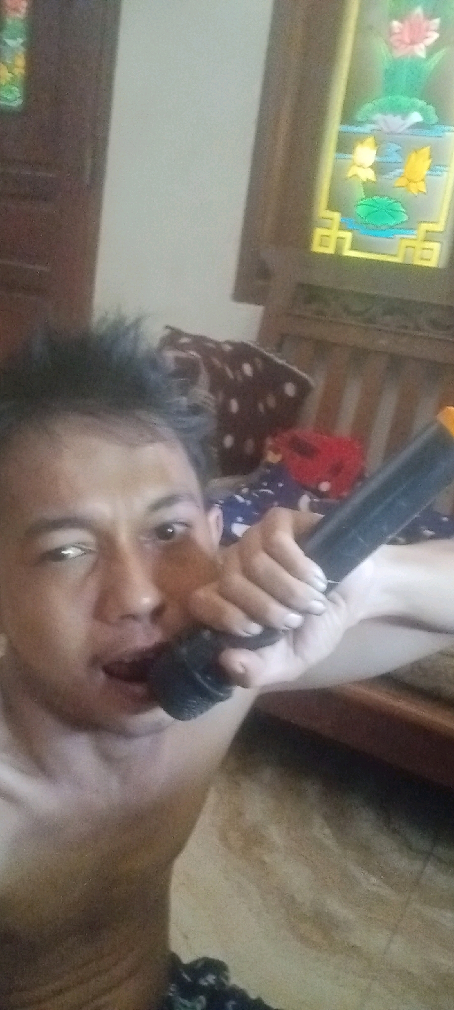 The image shows a close-up selfie of a shirtless man holding a microphone to his mouth. The man has short, spiky dark hair and is looking directly at the camera with his mouth slightly open. He is holding the microphone in his right hand, with his left hand near his face. The background appears to be an indoor setting with a wooden chair, some blankets, and a decorative window with floral imagery. The lighting is dim and casts shadows. The image quality is low, with visible grain and pixelation. The man's ethnicity appears to be Asian. Based on the provided information, the location is identified as Karawang, Indonesia.