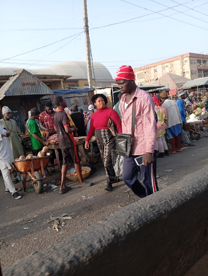 The image depicts an outdoor market scene in Sabon Gari West, Nigeria, under a clear, light blue sky. The ground is an unpaved or damaged asphalt surface littered with debris, including dry plant matter and refuse.
In the foreground, a concrete barrier runs horizontally across the bottom right. A male individual walks from right to left, wearing a red and white striped knit cap, a red and white plaid long-sleeved shirt, dark blue track pants with a red and white side stripe, and black shoes. He carries a black shoulder bag on his left side and holds a white smartphone in his right hand. His gaze is directed forward and slightly downward.
Mid-ground, a female individual stands to the left of the foreground male, wearing a red long-sleeved top and dark patterned trousers. Her dark hair is pulled back. To her left, a younger male, largely facing away, stands near a wooden market table, wearing a dark t-shirt with red accents and dark shorts. Next to him, a basket containing oranges or similar round fruit rests on the ground. Further left, a male in a light green tunic and dark pants stands beside an orange wheelbarrow filled with coconuts or large round fruits.
The background features numerous informal market stalls constructed from corrugated metal sheets and other makeshift materials, indicating extensive commercial activity. Various other individuals, appearing to be vendors and shoppers, are visible throughout the crowded market area, dressed in diverse casual attire. Some distant multi-story buildings and a large, dome-like structure are visible beyond the market stalls. A tall utility pole with overhead wires extends upwards from the left mid-ground.