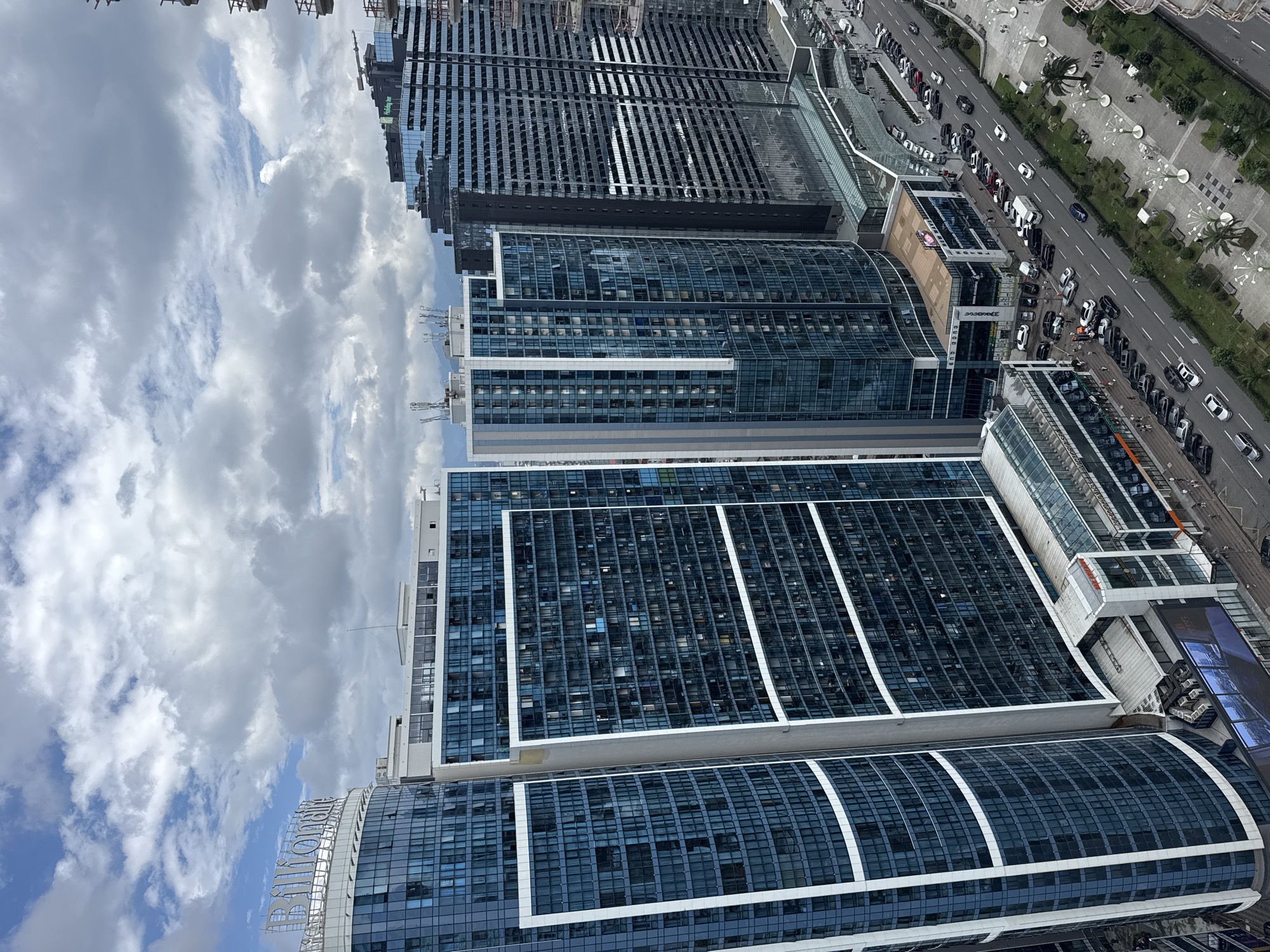 An aerial, high-angle shot captures a cityscape featuring multiple high-rise buildings with predominantly blue glass facades under a cloudy sky. The foreground showcases several of these modern skyscrapers, one with the illuminated sign "Billionaire" visible at its apex. Adjacent to these are other tall structures, including one with the "Holiday Inn" logo. A street with moderate traffic, including cars and a white van, runs alongside the buildings. Pedestrians are present on the sidewalks, and the area is landscaped with palm trees and decorative streetlights. The architecture suggests a modern urban environment, consistent with coastal developments in Batumi, Georgia.
