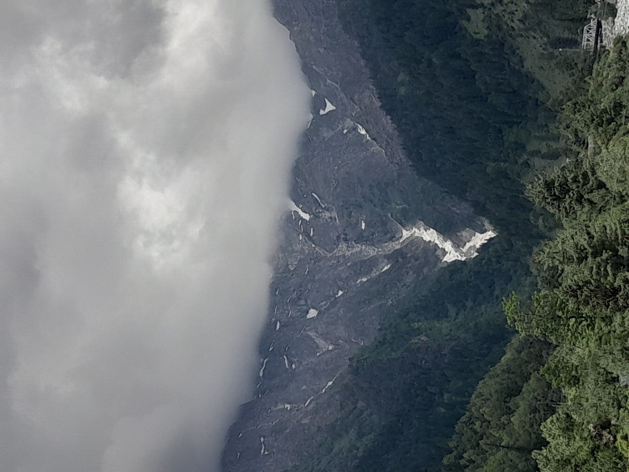 An overcast sky looms over a dramatic mountain landscape. The jagged, dark gray peaks are partially obscured by thick, billowing clouds that hang low, creating a moody and atmospheric scene. Patches of snow dot the upper slopes of the mountains, stark against the dark rock. A prominent V-shaped ravine cuts through the center of the mountains, filled with what appears to be a snowmelt or rocky stream, contrasting with the surrounding dark forest. The lower slopes are densely covered in dark green pine trees, forming a thick canopy that extends down into a valley. In the foreground, more trees, lighter in color and more varied in shape, create a lush border. On the far right, a small, white bridge with a truss structure is visible crossing a body of water, hinting at human presence in this otherwise wild environment. The overall impression is one of rugged natural beauty and a somewhat somber, cloudy day.