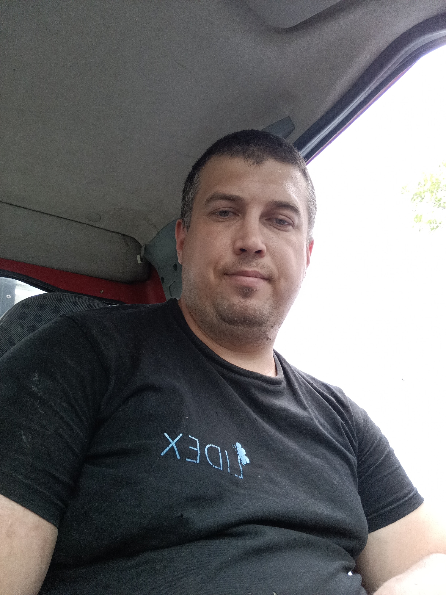 A man with short brown hair and a light beard is photographed from the chest up, sitting inside what appears to be a vehicle. He is wearing a black t-shirt with a light blue logo consisting of the letters "IDEX" and a stylized butterfly or flower. The man is looking directly at the camera with a neutral expression. The interior of the vehicle is visible, including the grey headliner and a red door panel. The background outside the vehicle is a bright, overexposed sky with hints of green foliage. The photograph was taken in Svémyslice, Czechia.