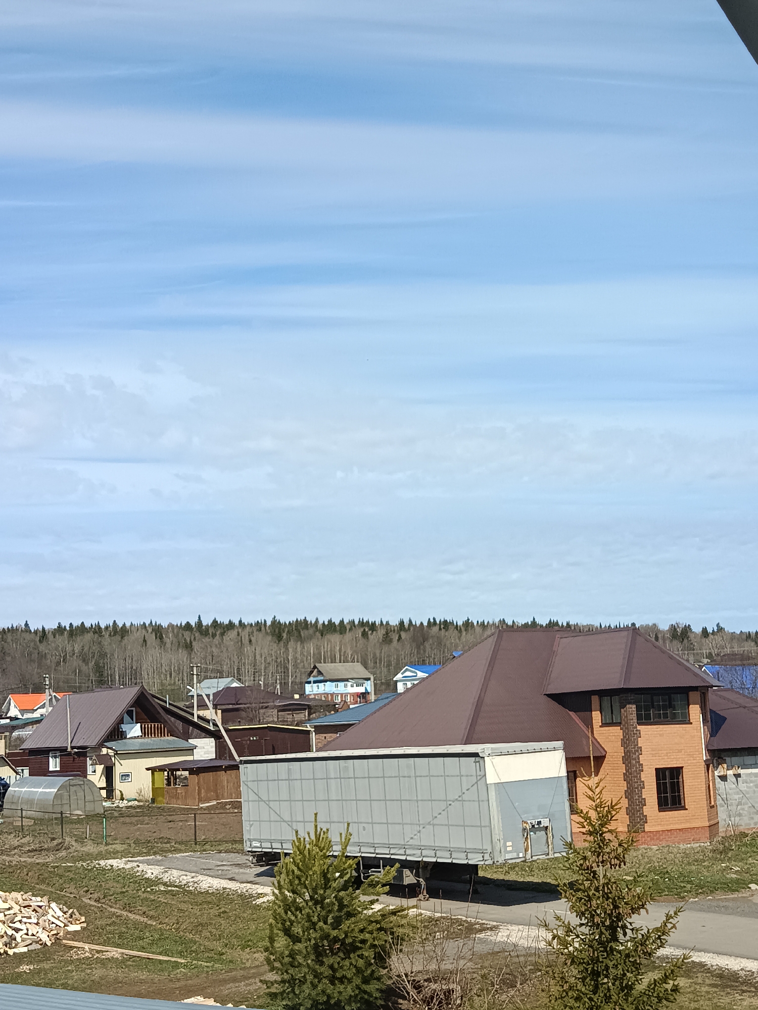 The image depicts a rural or suburban scene, potentially located in Barda, Russia, but without further information, the location cannot be definitively confirmed.

Foreground: Two small evergreen trees are present. A pile of split wood lies to the left.

Midground: A large gray trailer dominates the scene, situated on a dirt or gravel road. Behind the trailer, there are several residential buildings with varying architectural styles. One building is brick, with a brown roof and multiple windows. Other houses have wood siding and different roof colors. A small greenhouse is visible.

Background: A dense forest line stretches across the horizon, separating the built environment from the sky.

Sky: The sky is predominantly blue with thin, wispy clouds.

Overall: The scene suggests a residential area with a mix of older and newer construction. The presence of the large trailer implies potential industrial or commercial activity in the vicinity.