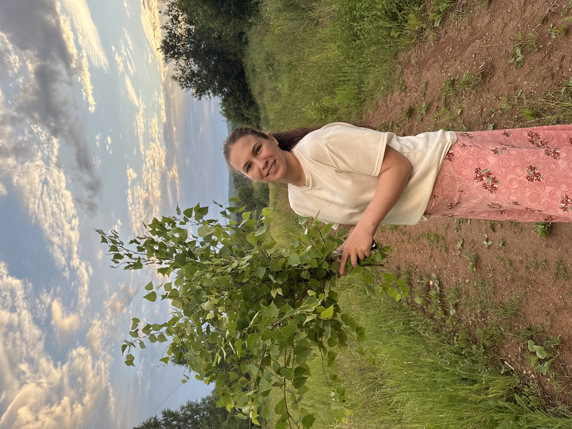 In the village of Nizhnyaya Gar', Russia, a young woman stands on a dirt path, holding a large bunch of green birch branches. She is wearing a white t-shirt and a pink skirt with a floral pattern. The background is a field of tall green grass and trees. The sky is a light blue with scattered clouds. The image appears to have been taken in the late afternoon or early evening.